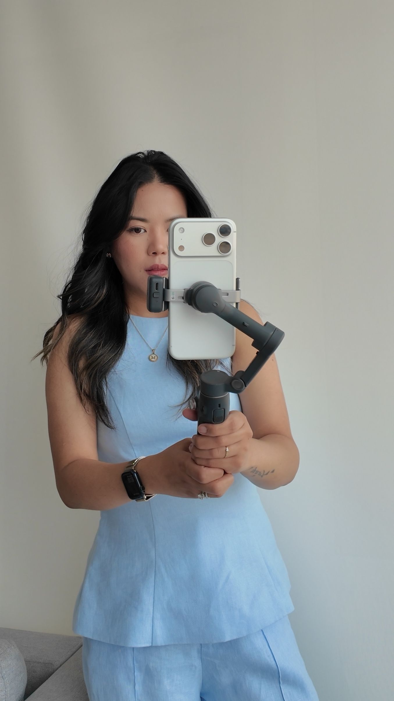 Woman in light blue outfit takes mirror selfie using smartphone mounted on a gimbal stabilizer.