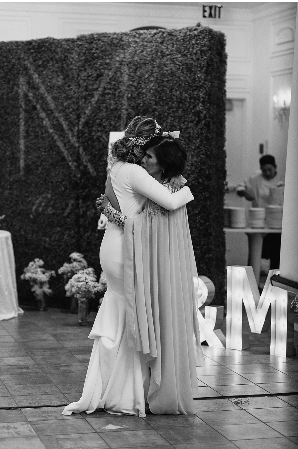 Bride and guest share emotional hug on dance floor before ivy wall backdrop at elegant wedding reception.