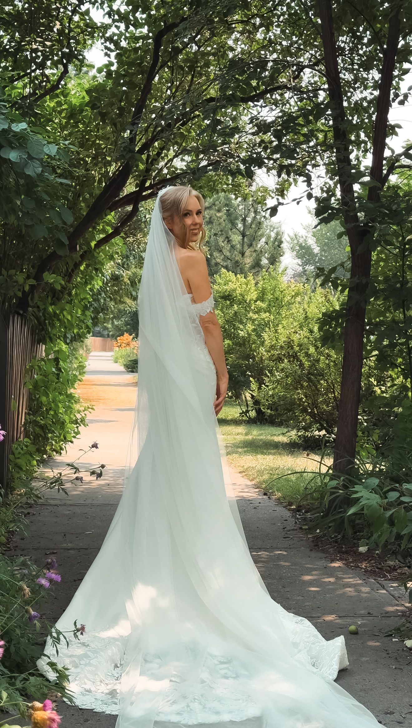 Elegant bride in off-shoulder lace gown with long veil poses on a sunlit garden pathway.