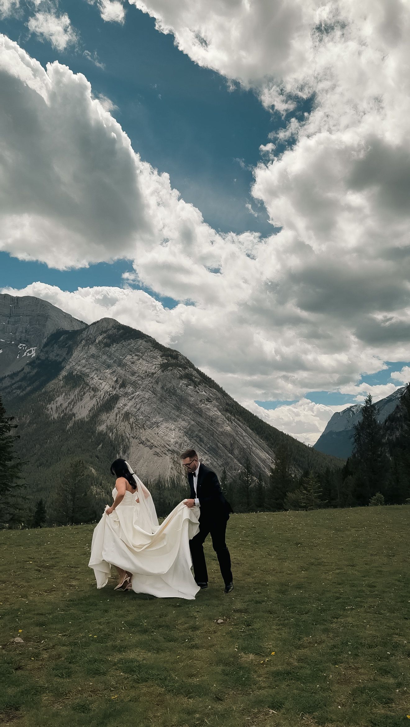 Bride and groom walking together on green meadow with stunning Rocky Mountain and cloudy sky background.