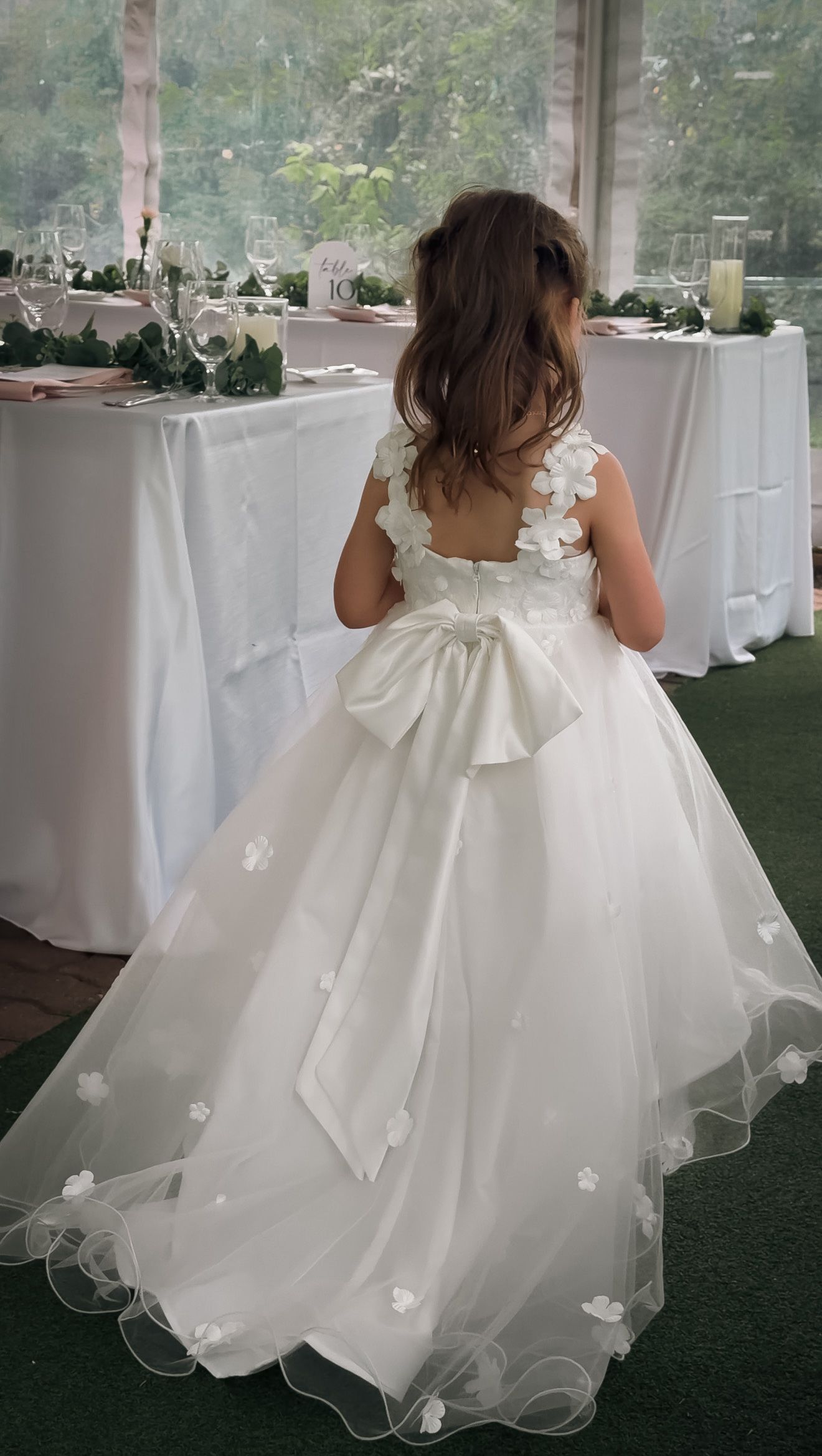 Young flower girl in white tulle gown with large bow standing at decorated wedding reception table.