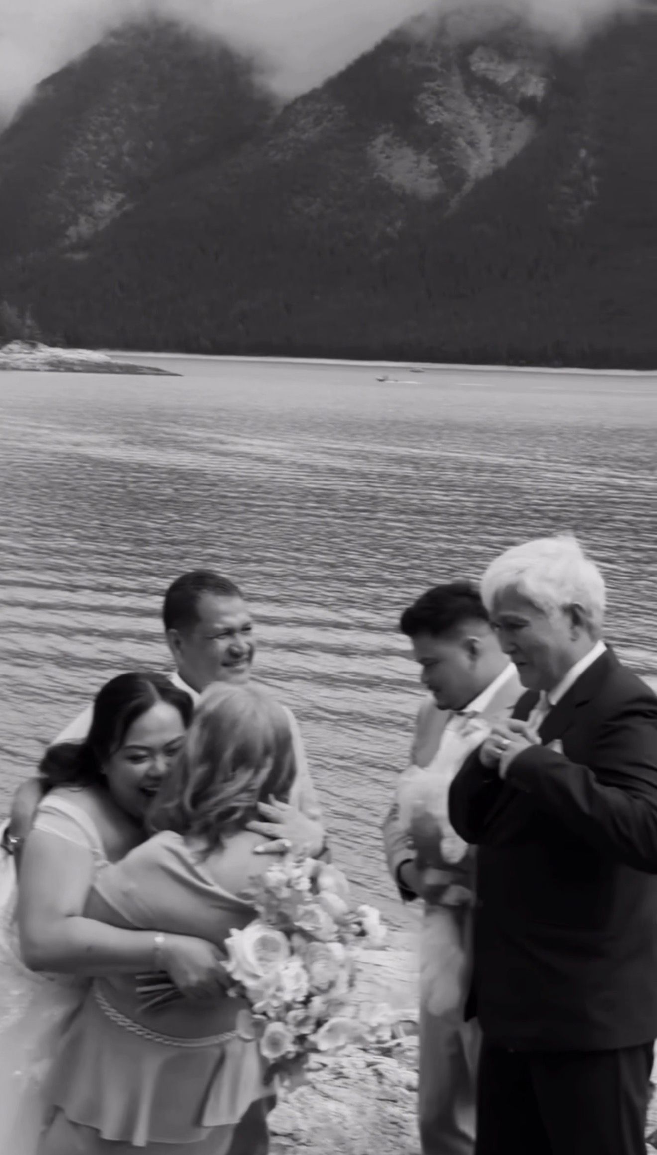 Black and white photo of wedding guests embracing and celebrating after an outdoor lakeside ceremony with mountains.