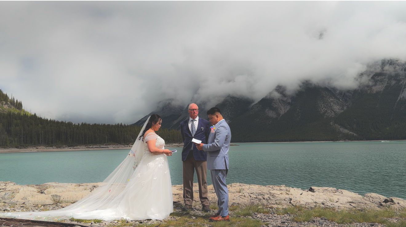 Outdoor wedding ceremony by a turquoise mountain lake with bride, groom, and officiant under cloudy skies.