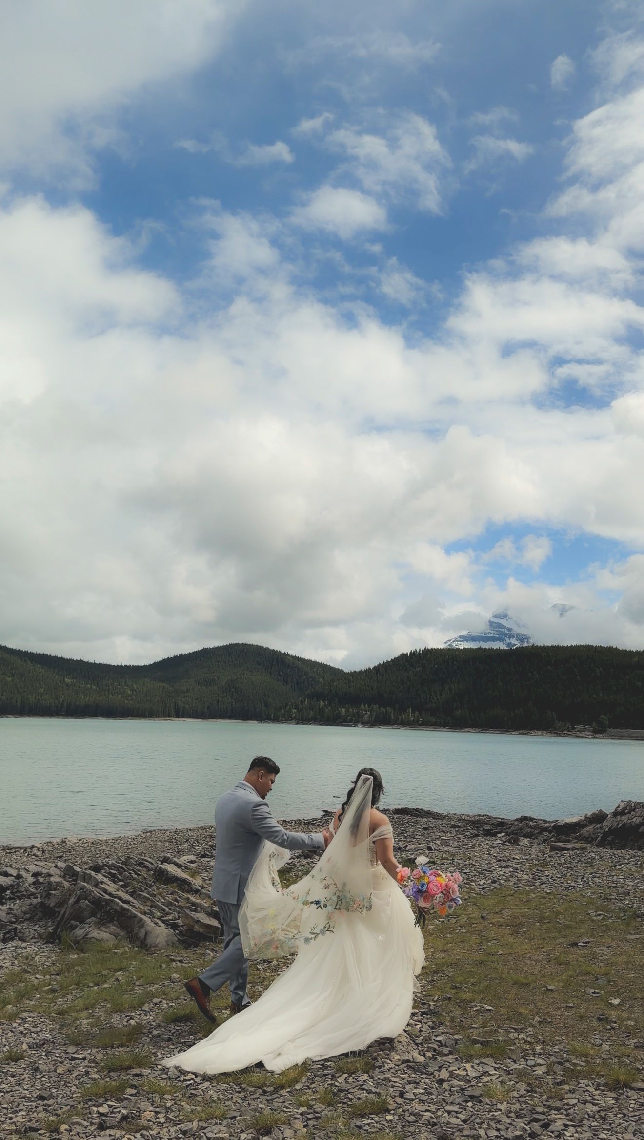 Bride and groom walking along rocky shoreline of a scenic mountain lake with colorful bouquet and flowing veil.