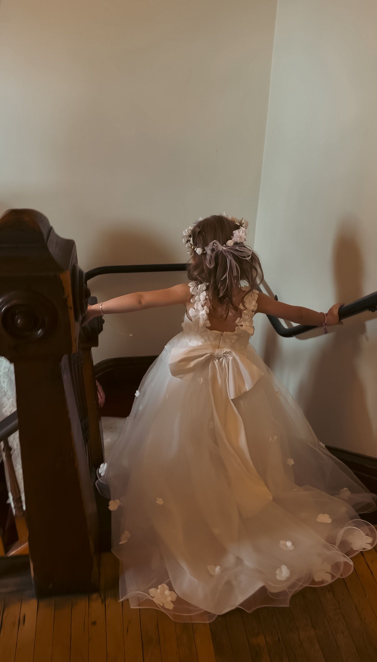 Young flower girl in a white tulle dress with floral hair accessories climbing a wooden staircase.