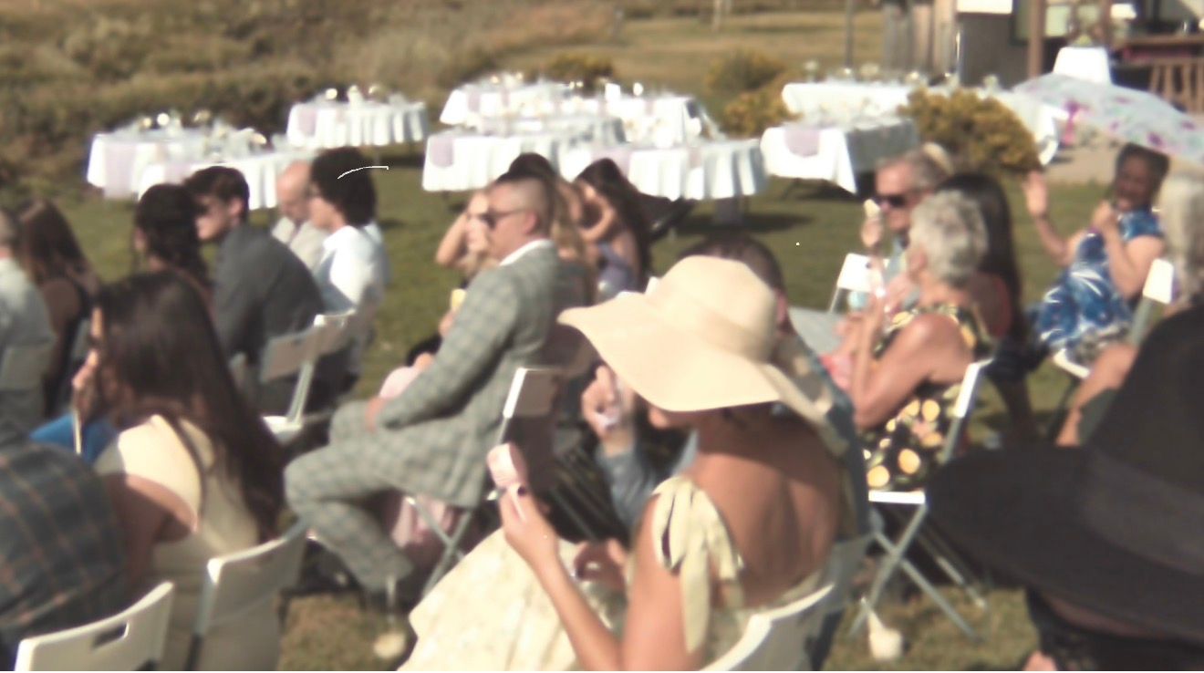 Outdoor wedding ceremony guests seated on white chairs in sunny field with round tables.