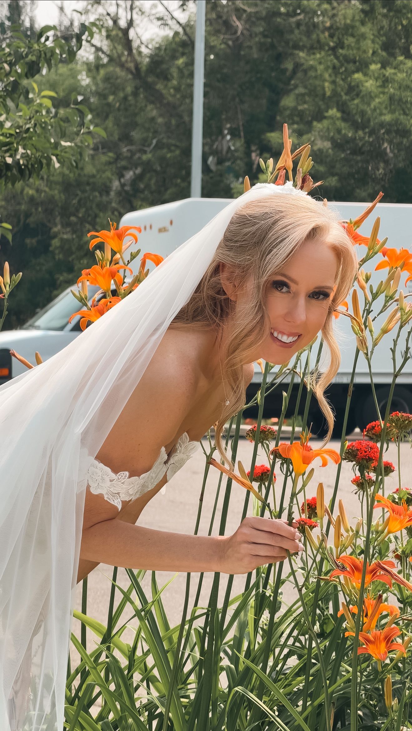 Smiling blonde bride in lace wedding dress and veil poses beside vibrant orange lilies outdoors.