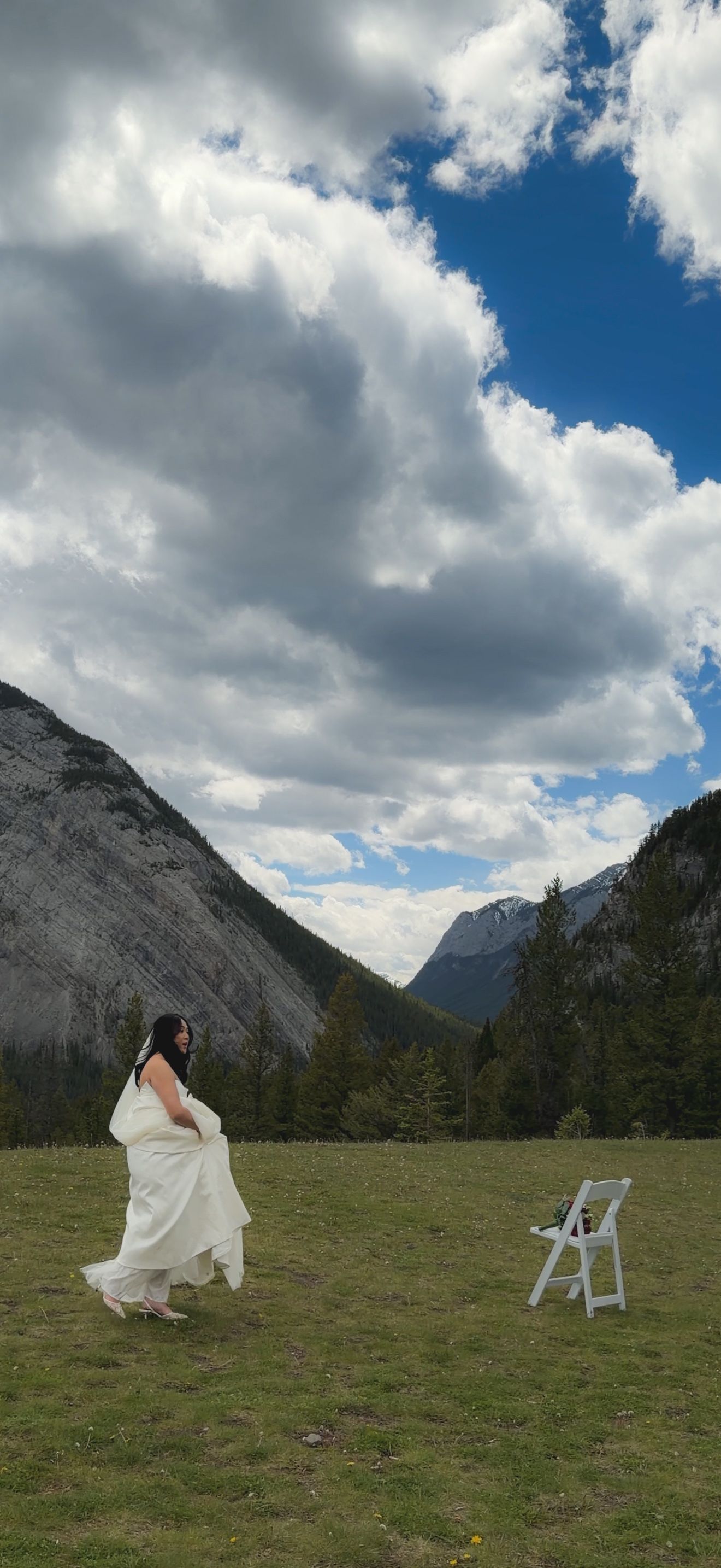 Bride in white wedding gown walking across green meadow with dramatic mountain and cloudy sky backdrop.