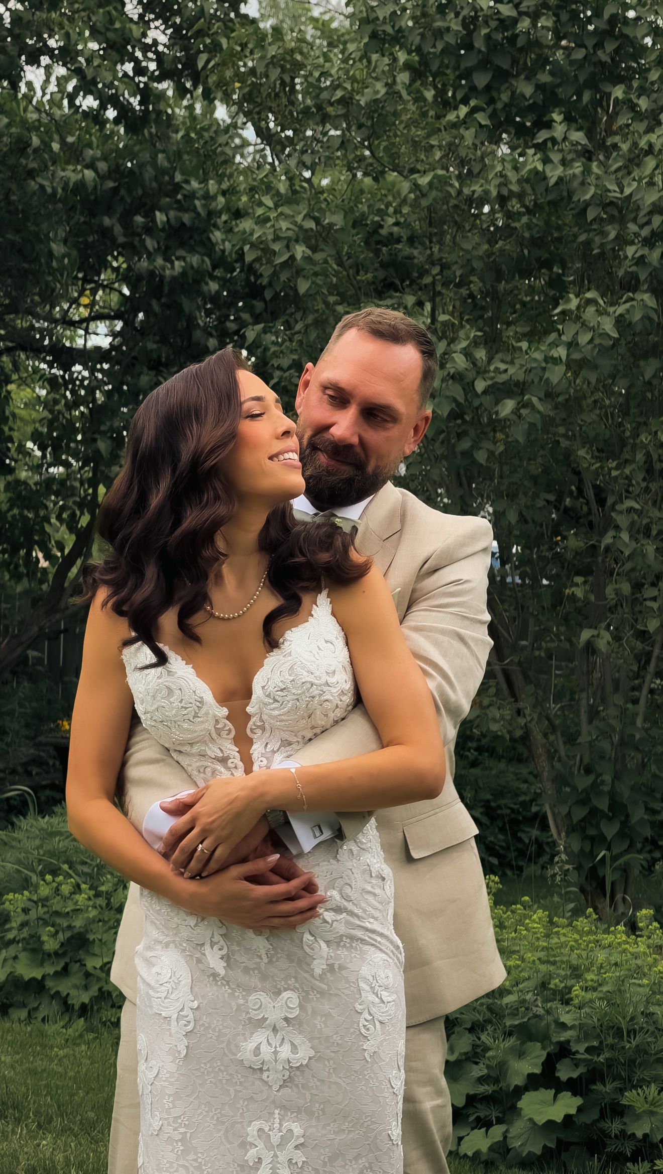 Groom in tan suit embracing smiling bride in lace wedding dress outdoors among green trees.