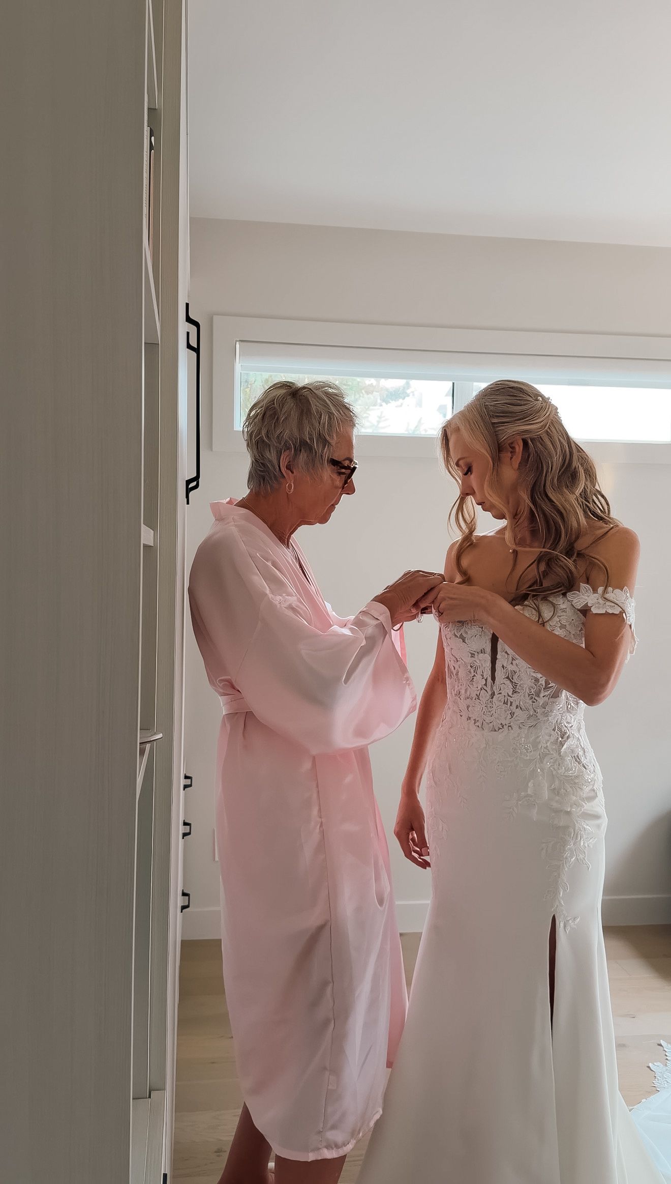 Older woman in pink robe helps blonde bride fasten her off-shoulder lace wedding gown.