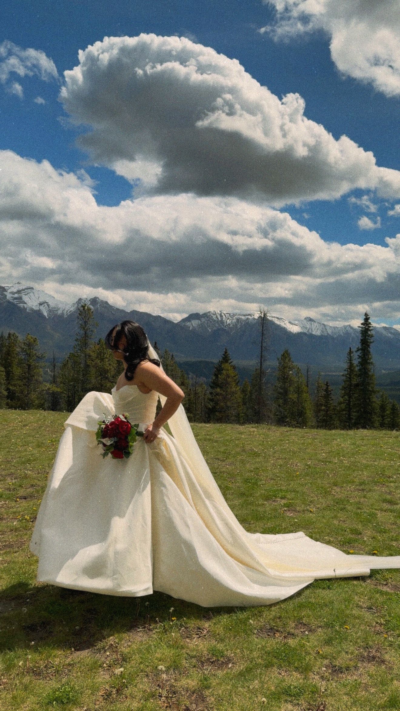 Bride in elegant ivory ball gown with long train poses with red bouquet against mountain landscape.