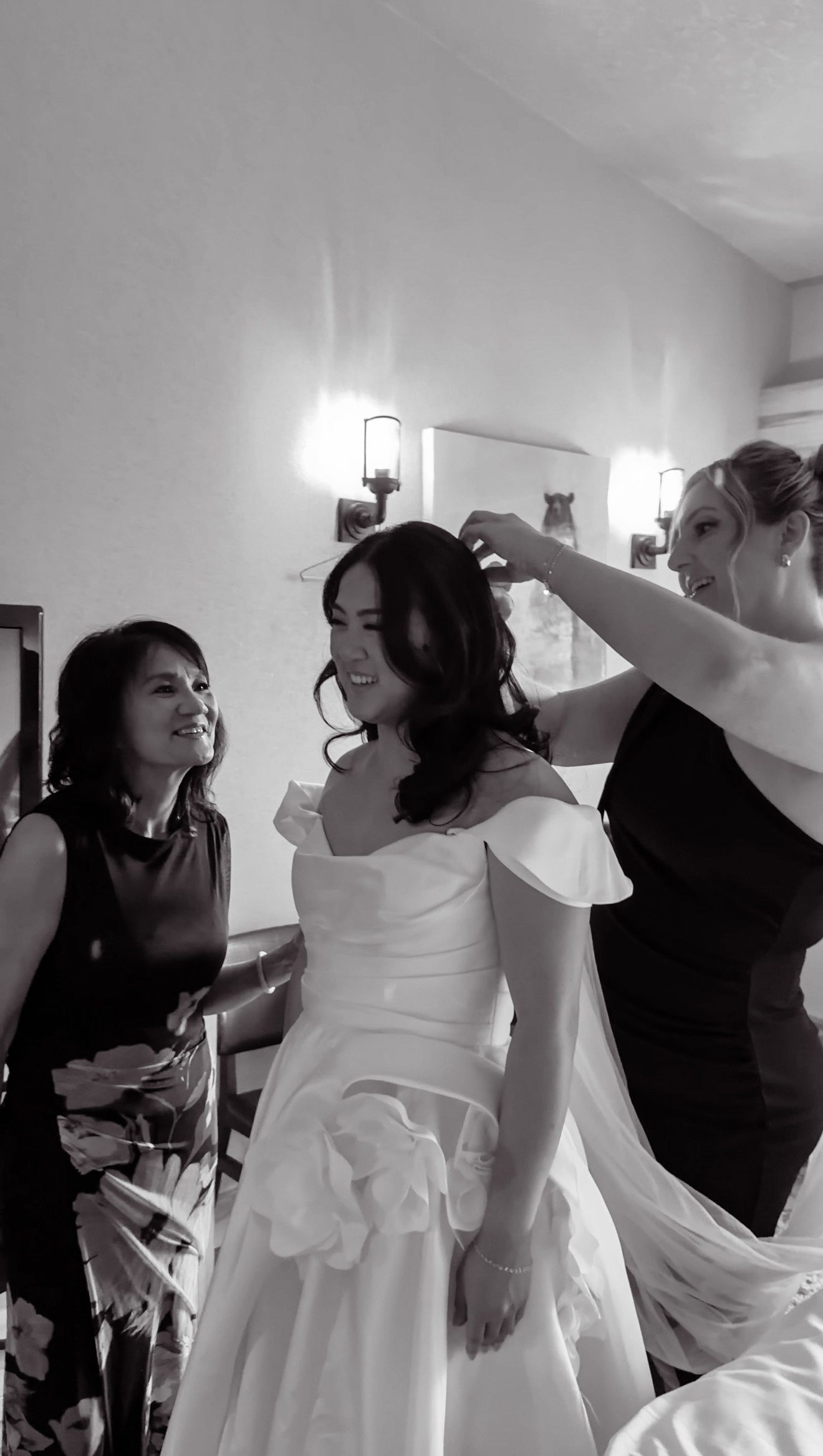 Black and white photo of bride smiling as attendants help with her veil and off-shoulder wedding gown.
