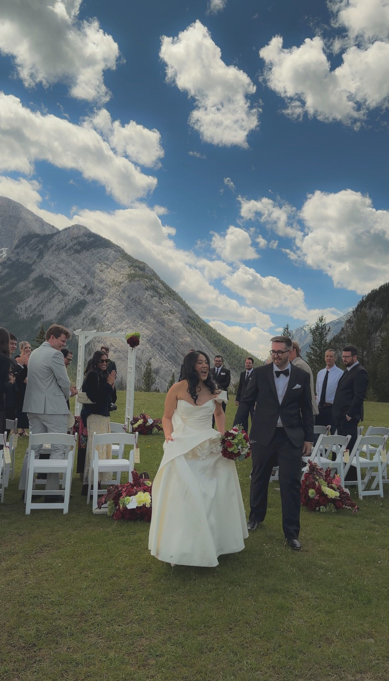 Laughing bride and groom recess down aisle at outdoor mountain wedding with white chairs and guests.