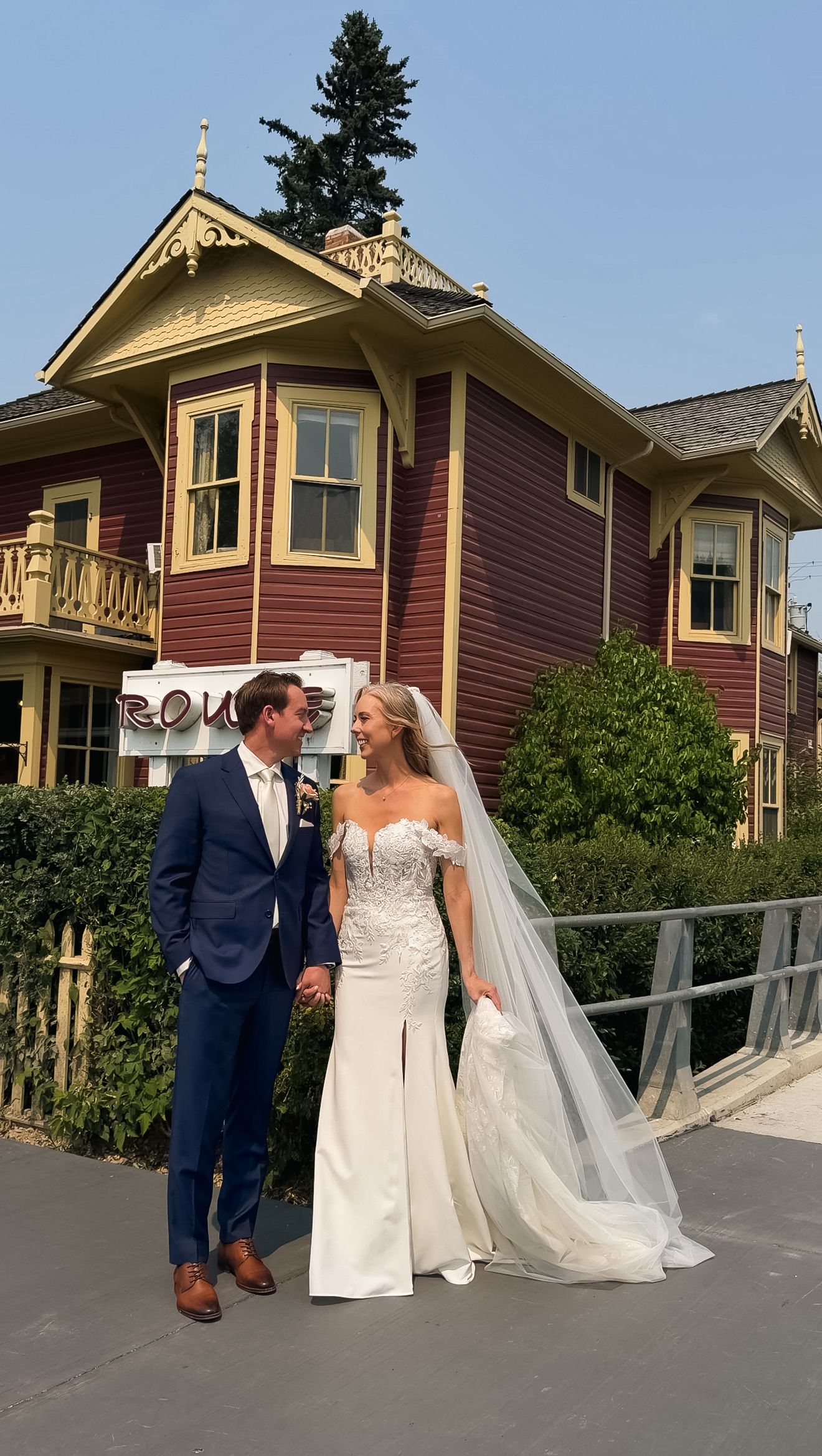 Bride in fitted lace wedding gown and groom in blue suit posing before a Victorian purple mansion.