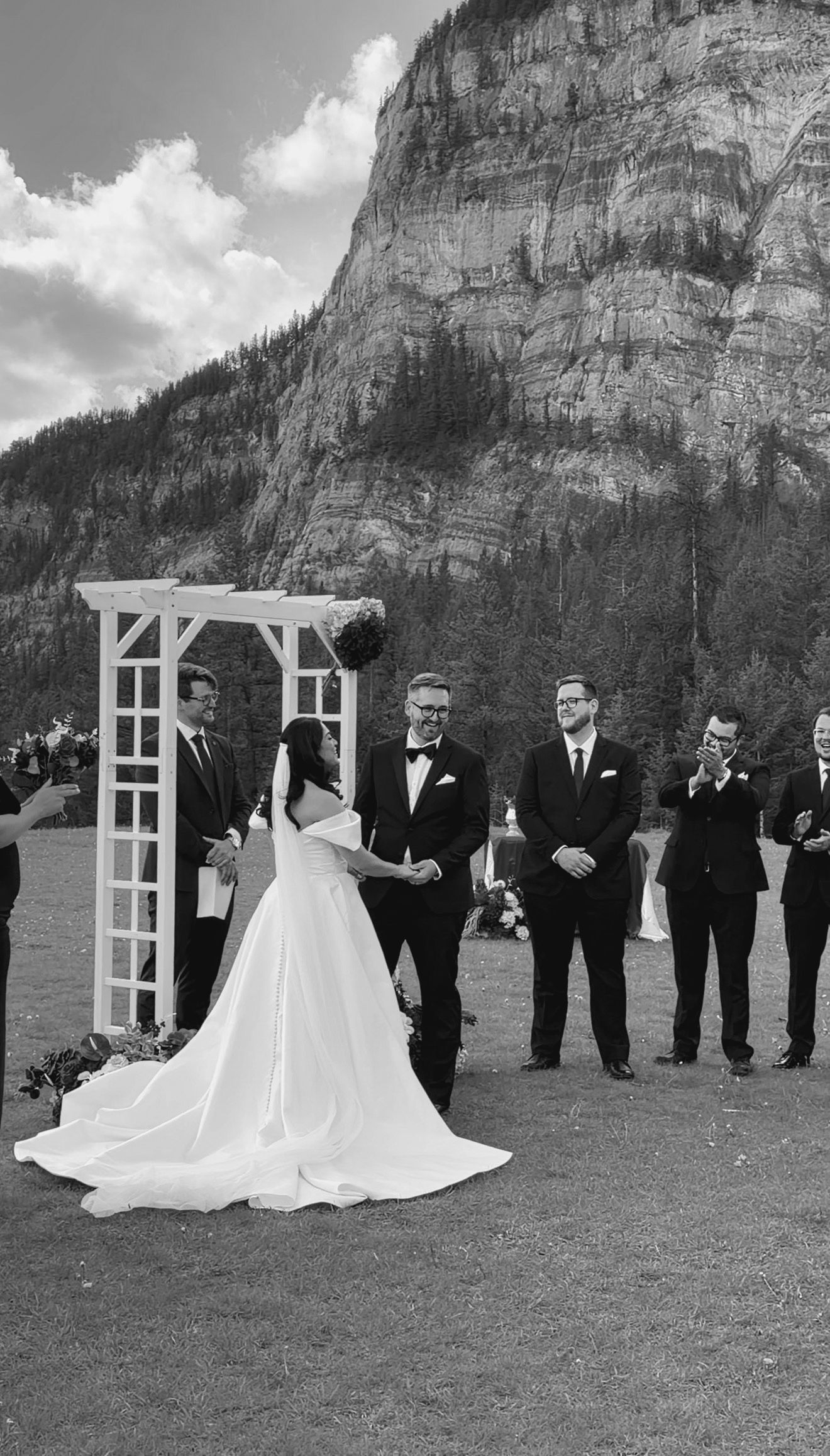 Black and white photo of bride and groom exchanging vows at outdoor mountain wedding with groomsmen watching.