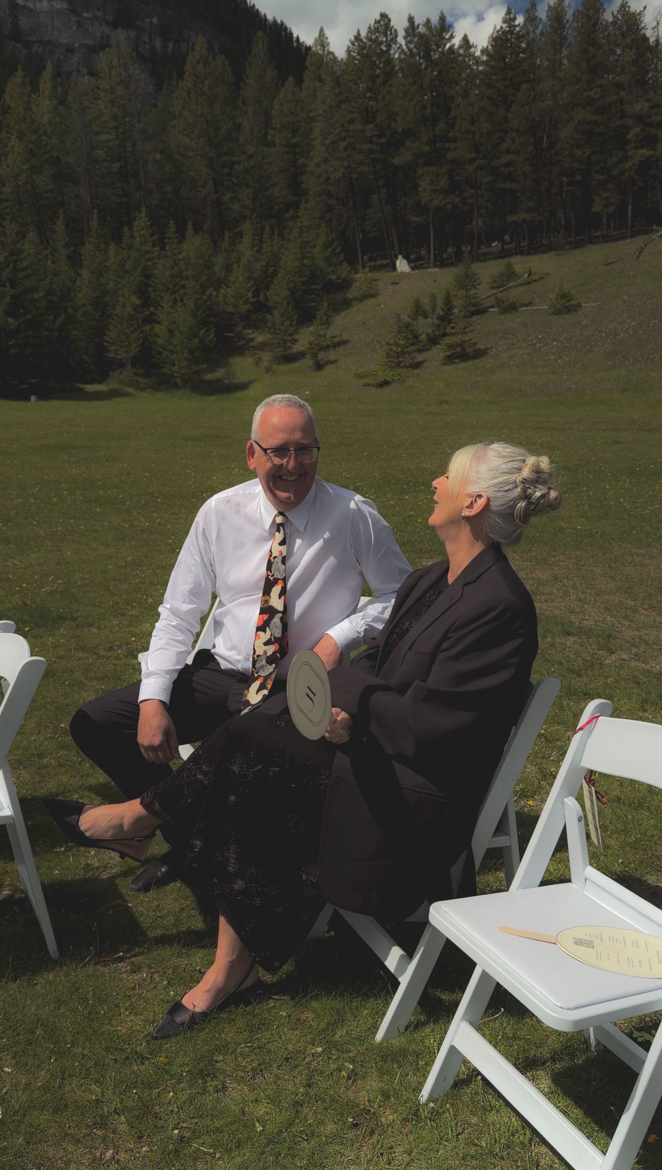 Couple sharing a joyful moment at outdoor mountain wedding seated in white chairs on green grass.