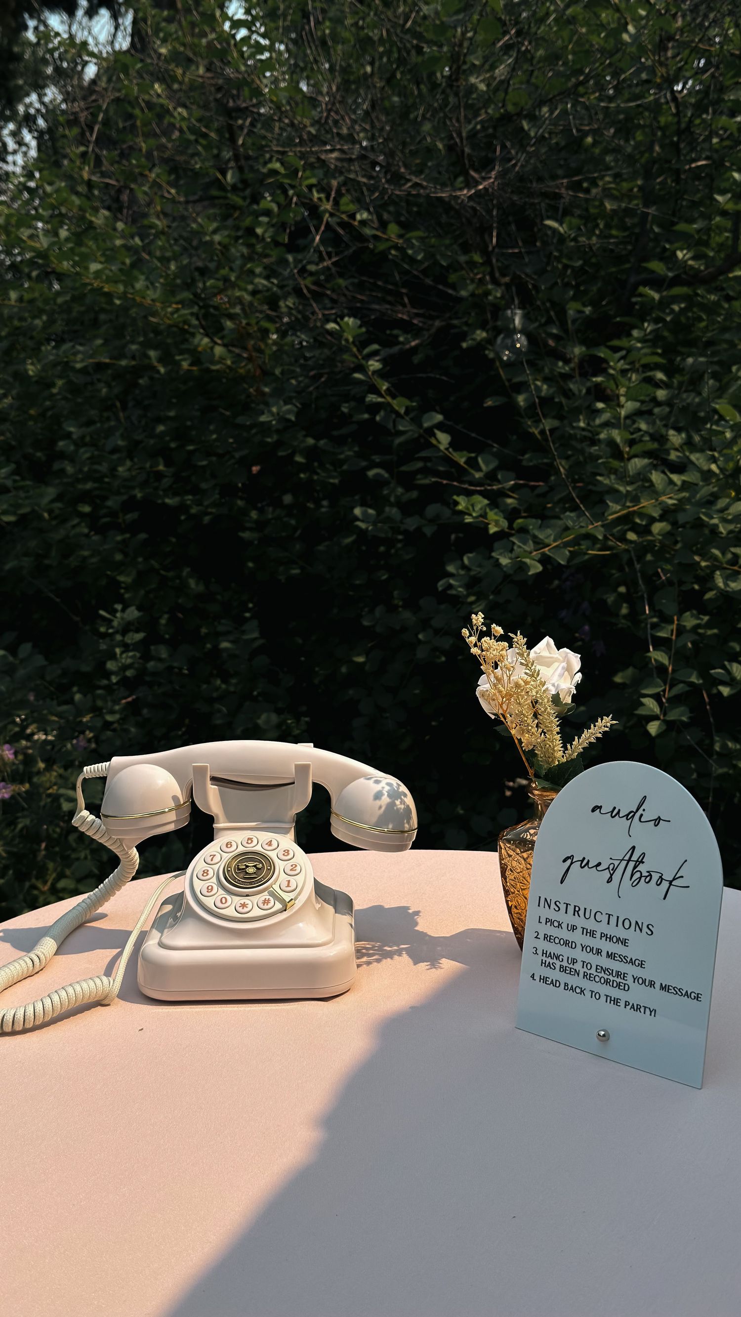 Vintage white rotary telephone audio guestbook display with floral arrangement at outdoor wedding.