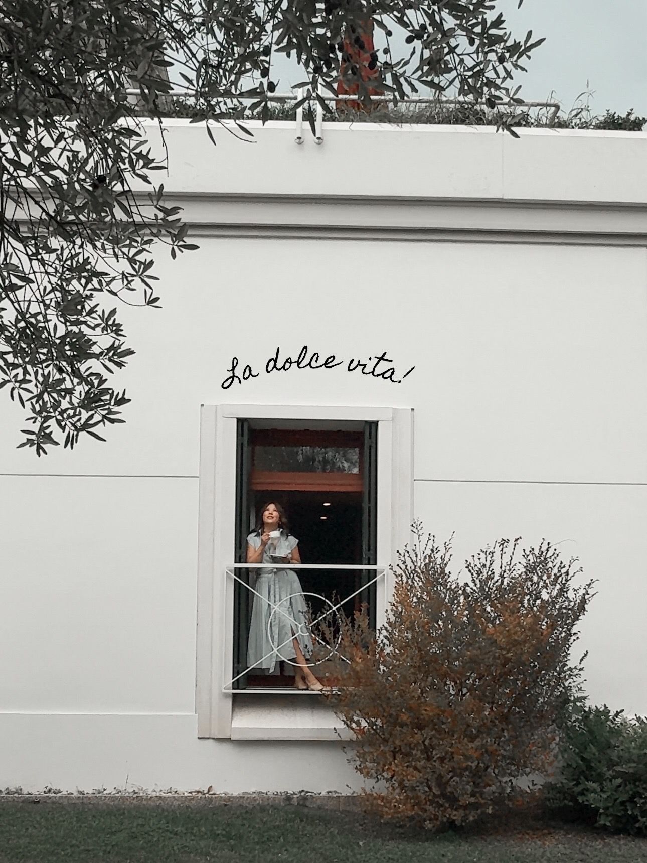 Woman in floral dress stands in window of white building with La Dolce Vita script written above.