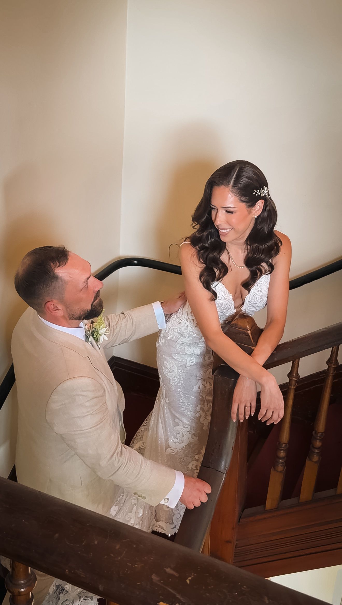 Joyful bride in lace gown laughs with groom in tan suit on elegant wooden staircase.