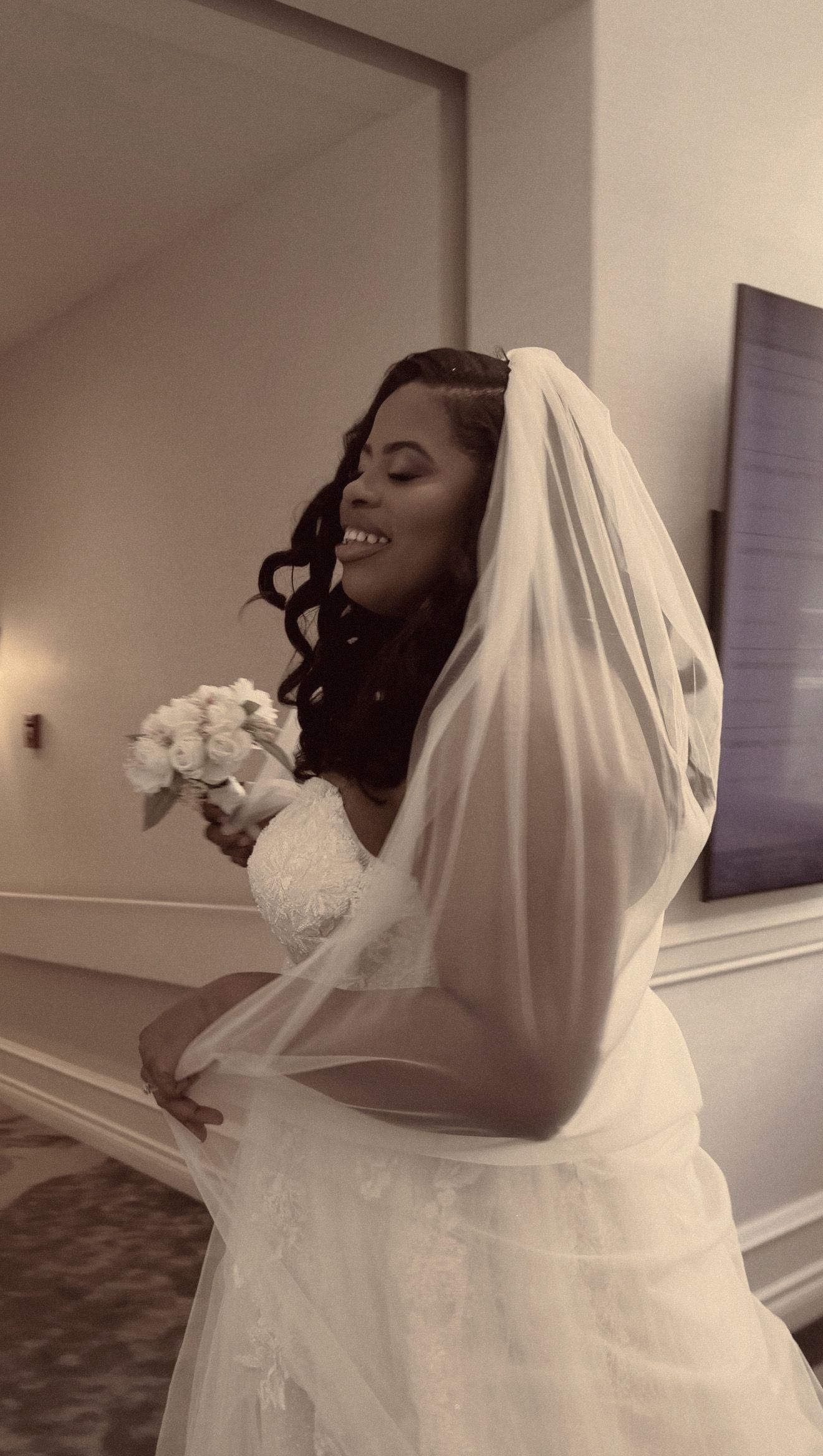 Beautiful Black bride smiles joyfully wearing a white veil and lace gown while holding a bouquet.