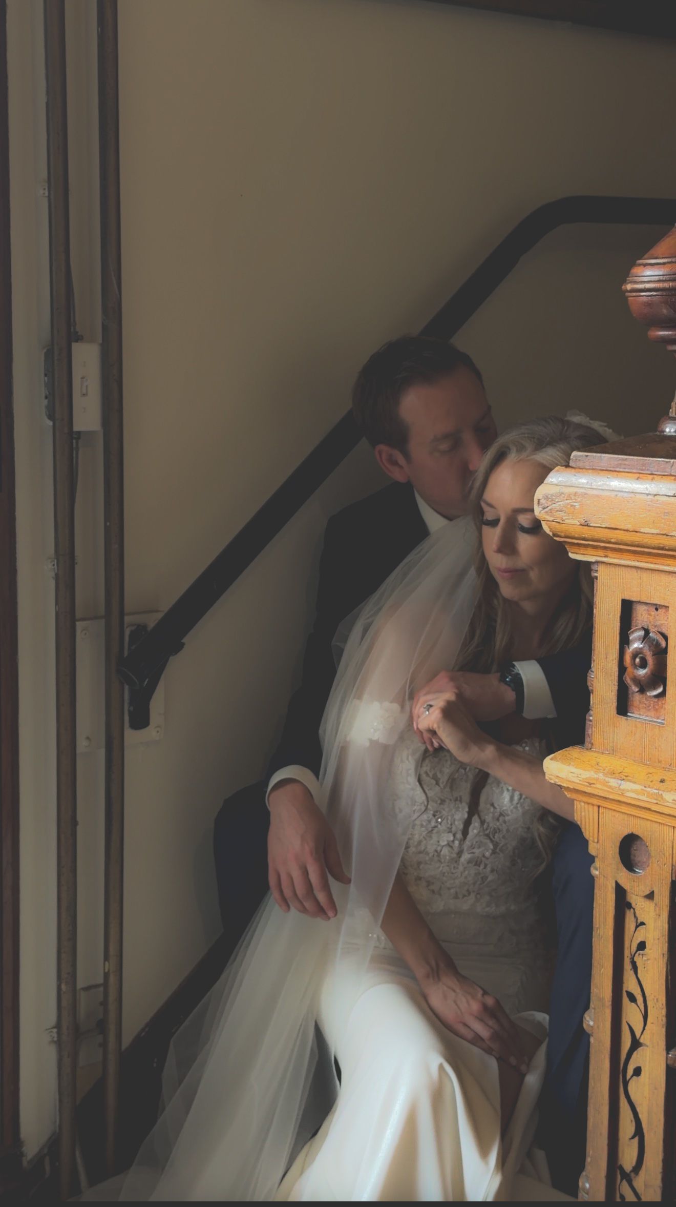 Groom tenderly kisses bride wearing veil and lace gown on decorative staircase.