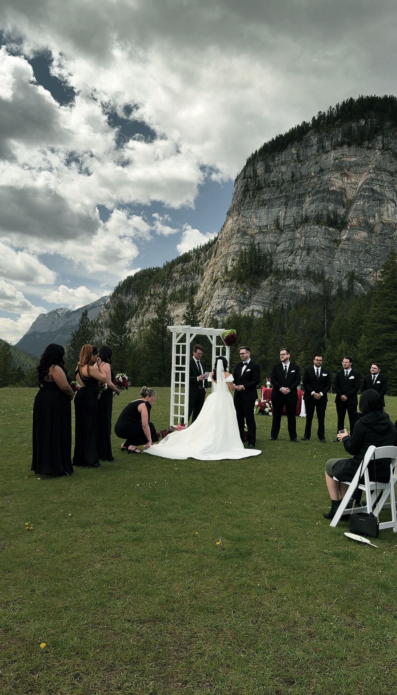 Outdoor mountain wedding ceremony with bride, groom, bridesmaids in black, and groomsmen under white arch in Banff.