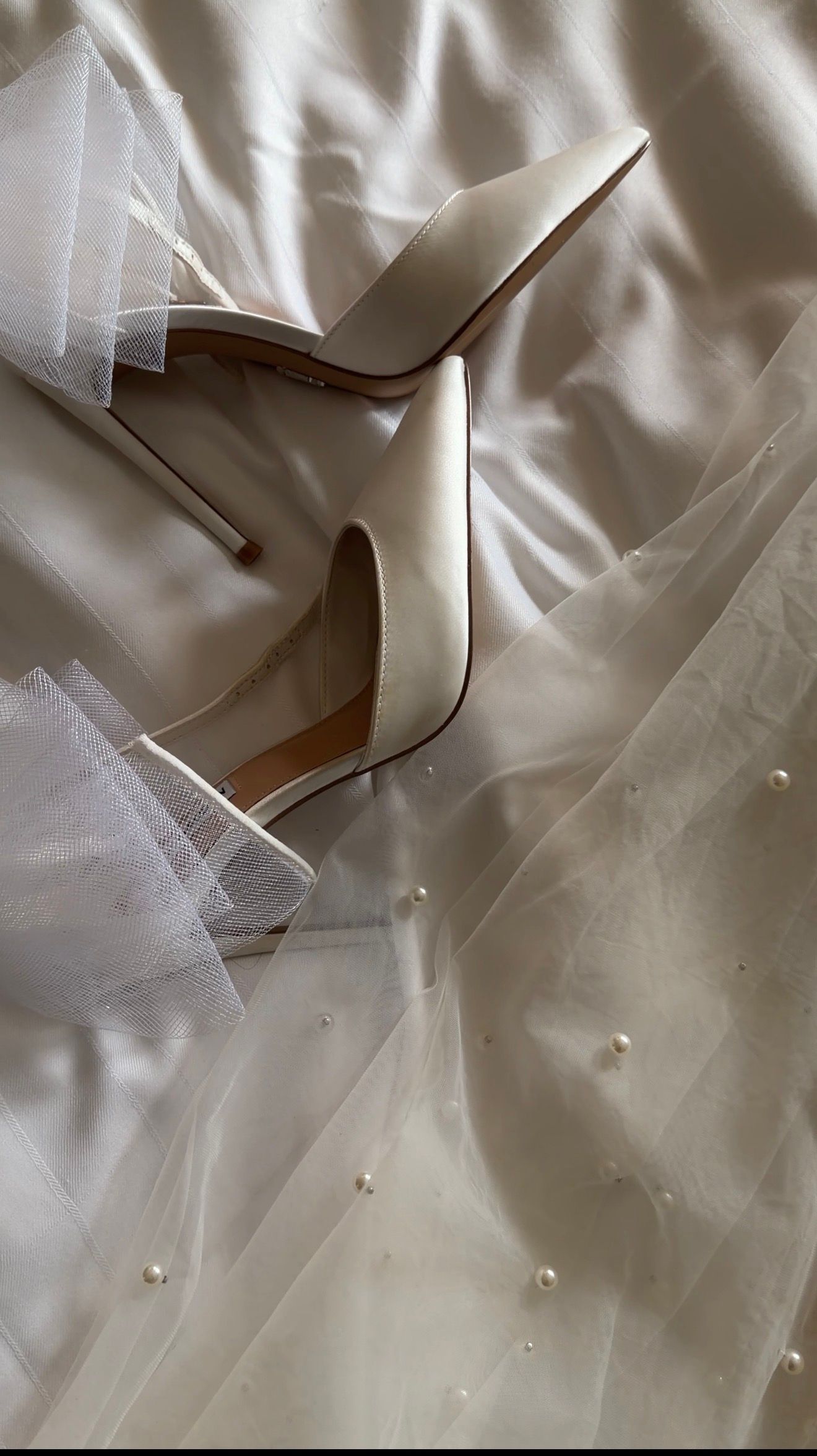 Elegant ivory bridal heels resting on a pearl-embellished wedding veil and satin dress on white bedding.