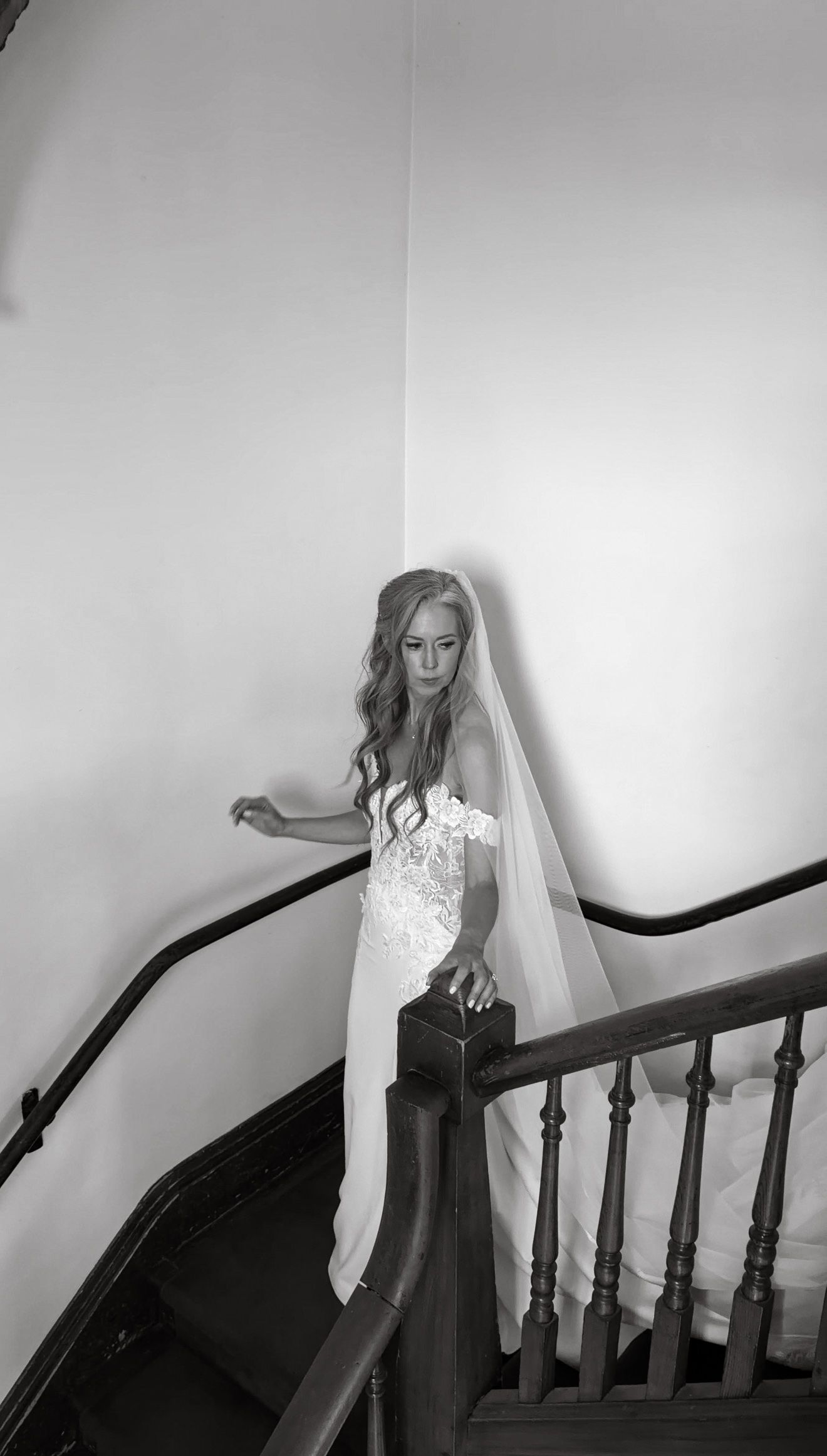 Elegant bride in lace off-shoulder wedding gown and veil descending a curved wooden staircase in black and white.