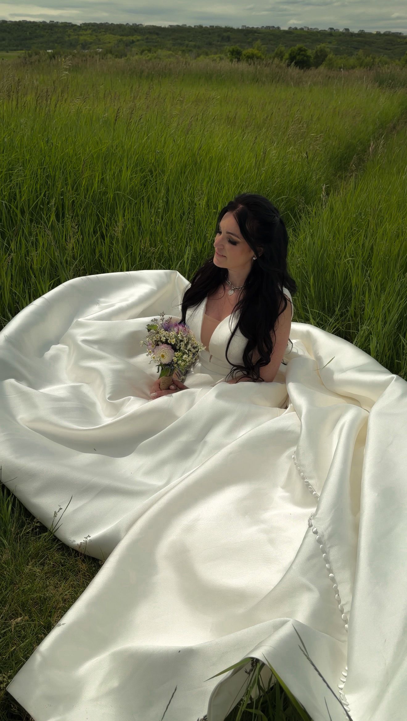 Dark-haired bride in white satin ballgown sitting in lush green field holding wildflower bouquet.