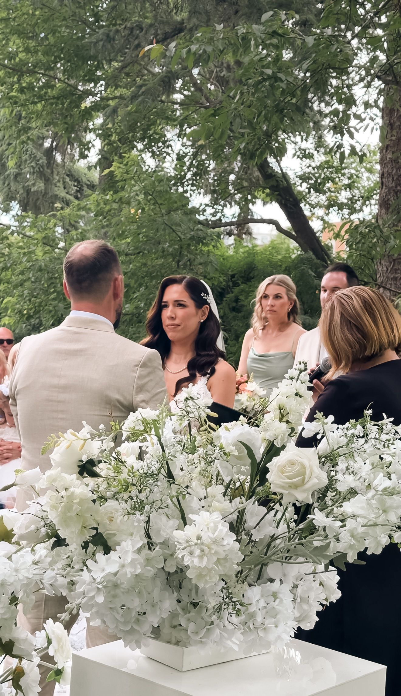 Bride exchanging vows at outdoor wedding ceremony surrounded by white floral arrangements and wedding guests.
