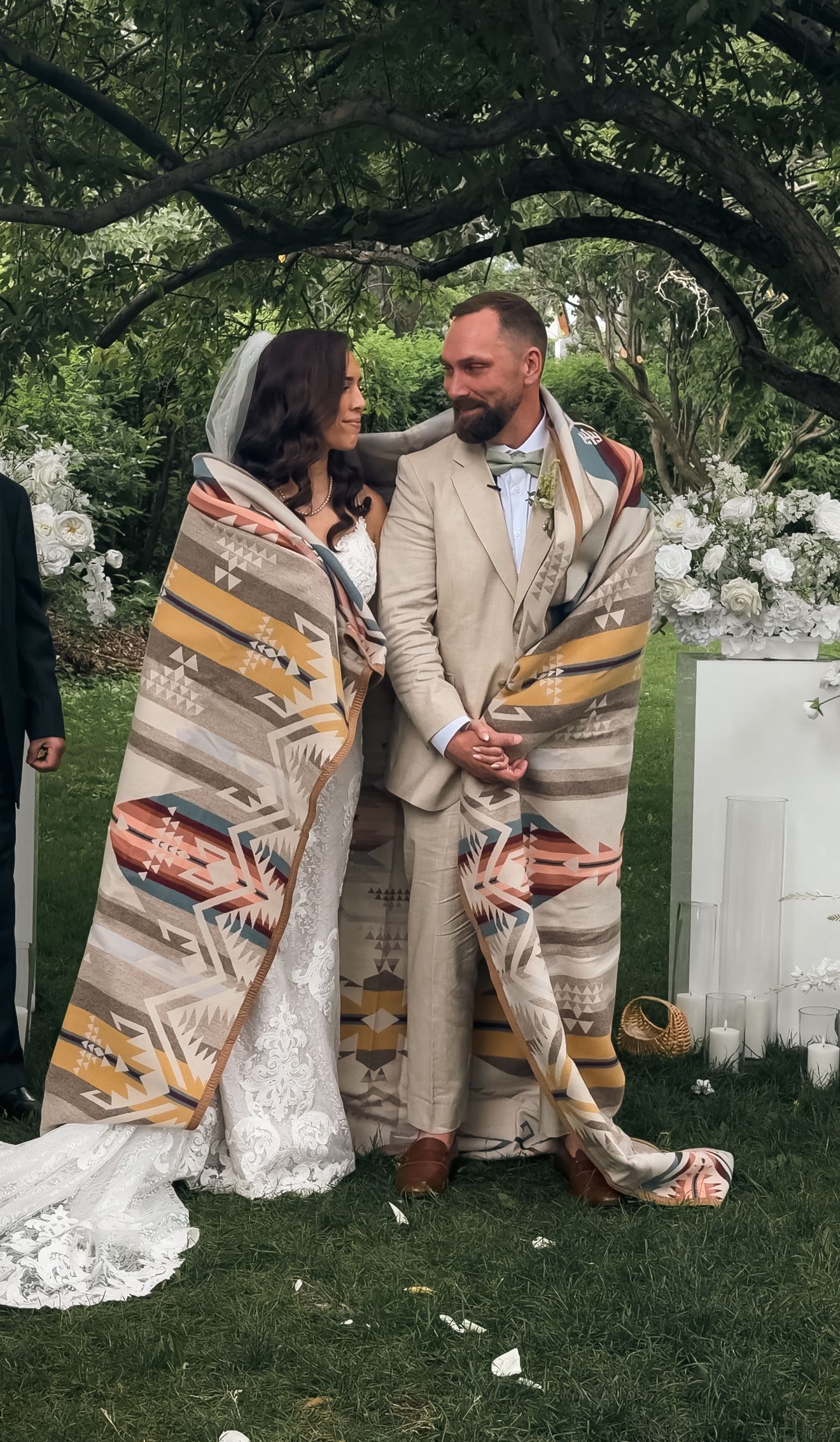 Bride and groom wrapped in a Pendleton blanket during an outdoor wedding ceremony surrounded by lush greenery.