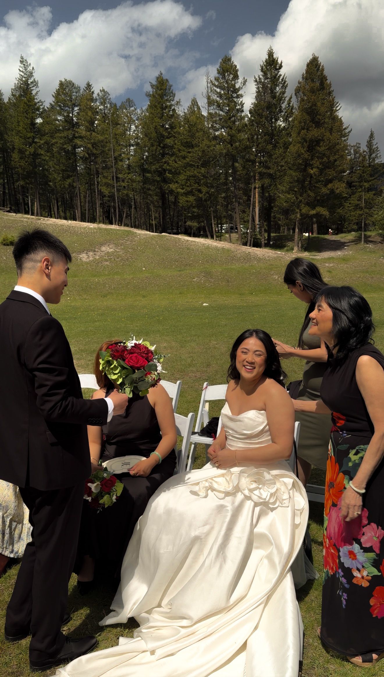 Smiling bride in white strapless gown holding red bouquet surrounded by guests at outdoor wedding.
