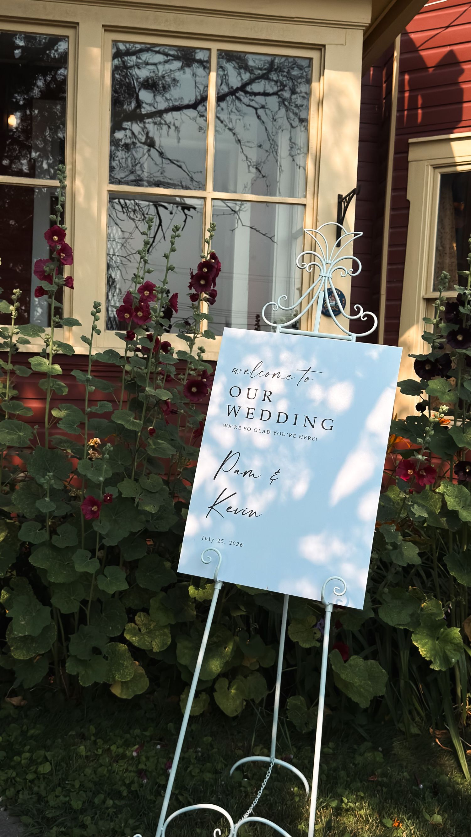 Welcome to Our Wedding sign for Pam and Kevin on a white easel surrounded by red hollyhock flowers outside a red house.