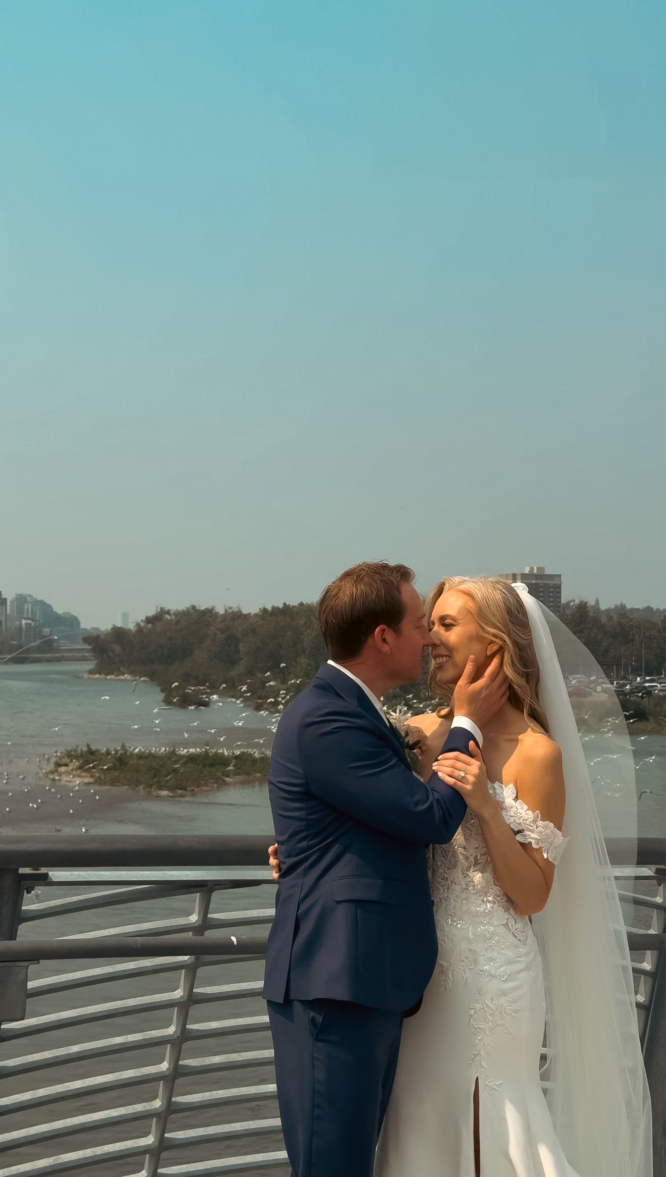 Groom in navy suit tenderly holds bride's face for a kiss on a waterfront bridge.