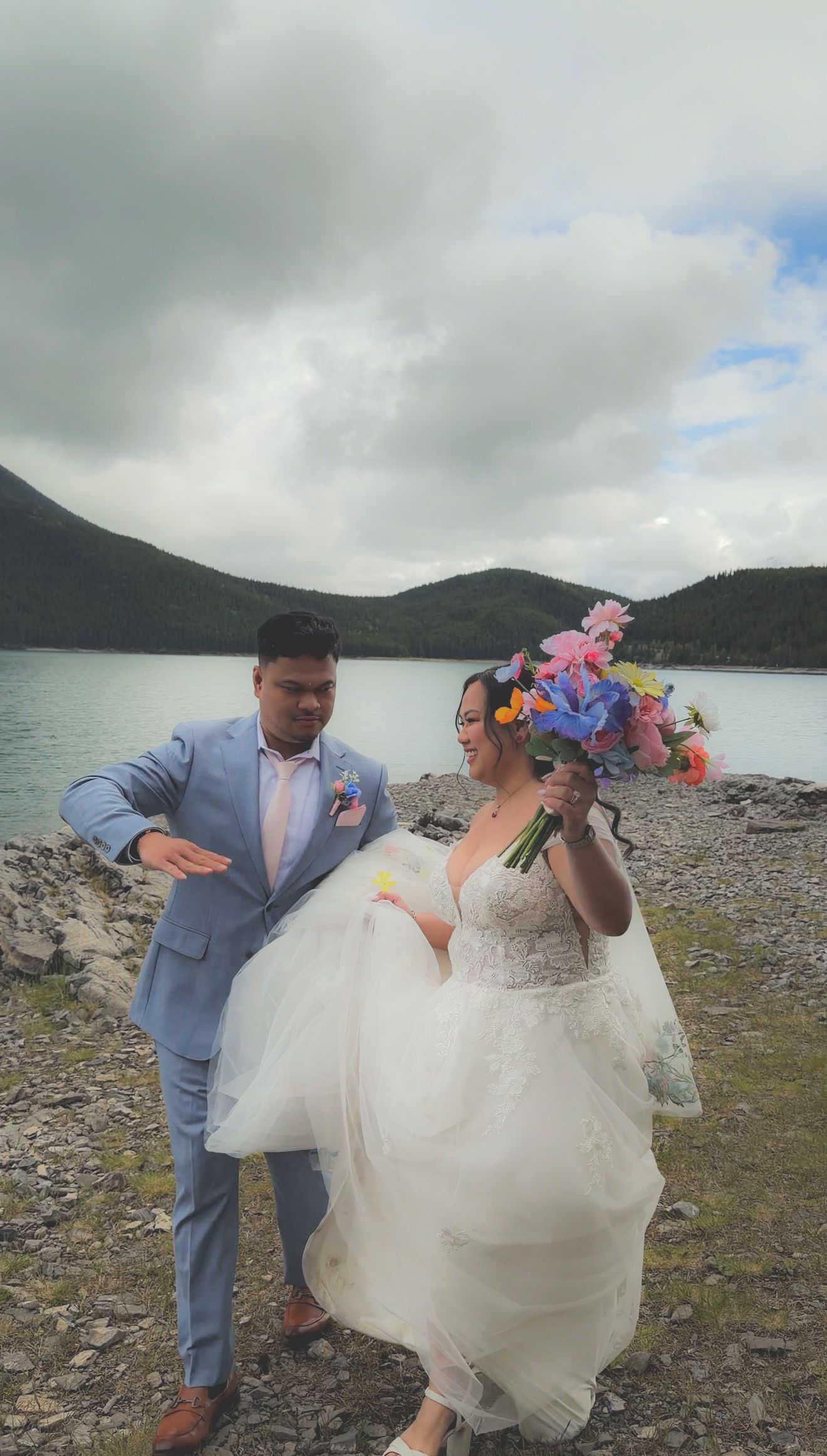 Bride and groom laughing together by a mountain lake, bride holding vibrant colorful floral bouquet in white gown.