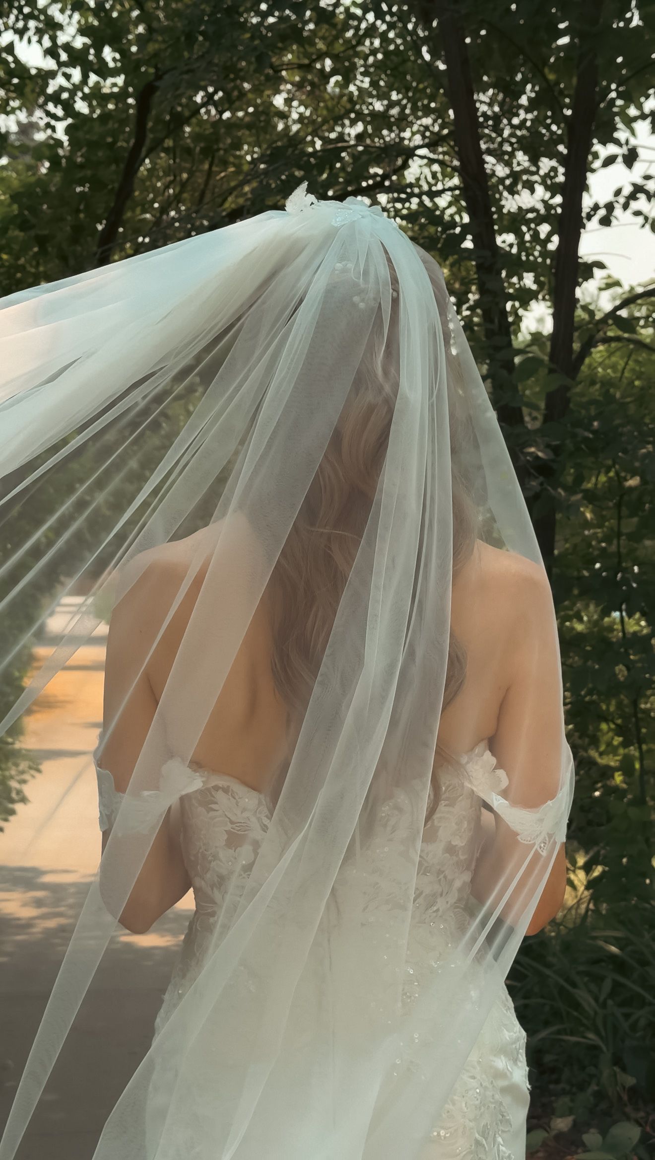 Bride in off-shoulder lace wedding gown with flowing veil stands among lush green garden trees.