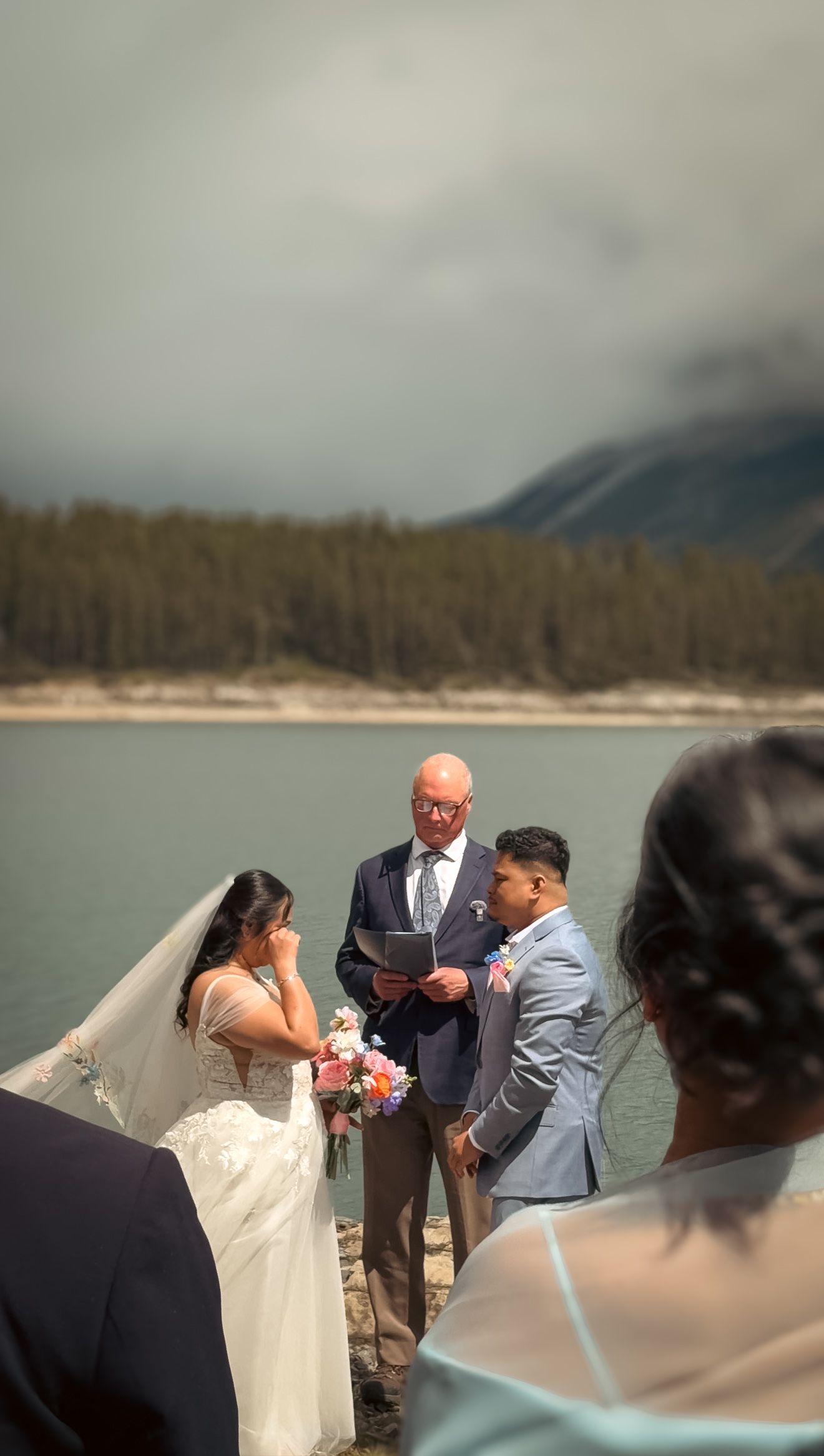 Emotional bride wiping tears during outdoor lakeside wedding ceremony with officiant and groom in light blue suit.