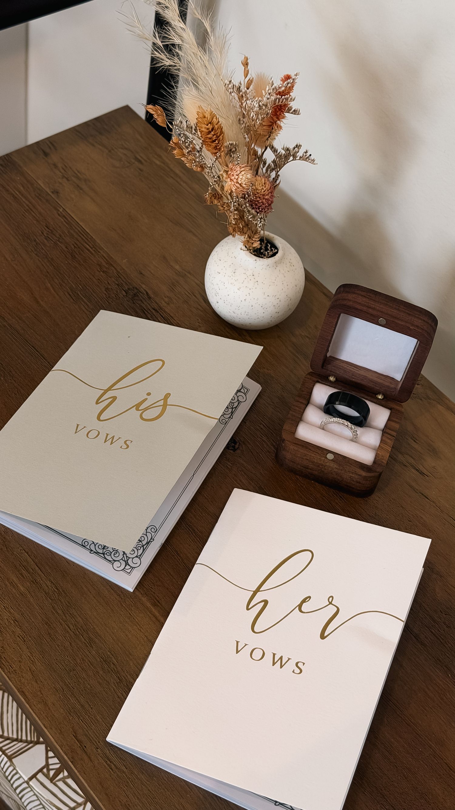 His and her vows booklets displayed on wooden table with a ring box and dried floral arrangement.
