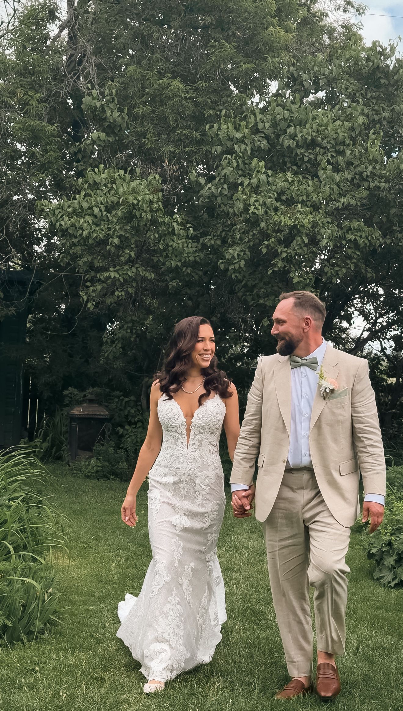 Bride in fitted lace gown and groom in tan suit walk hand in hand through lush green garden.