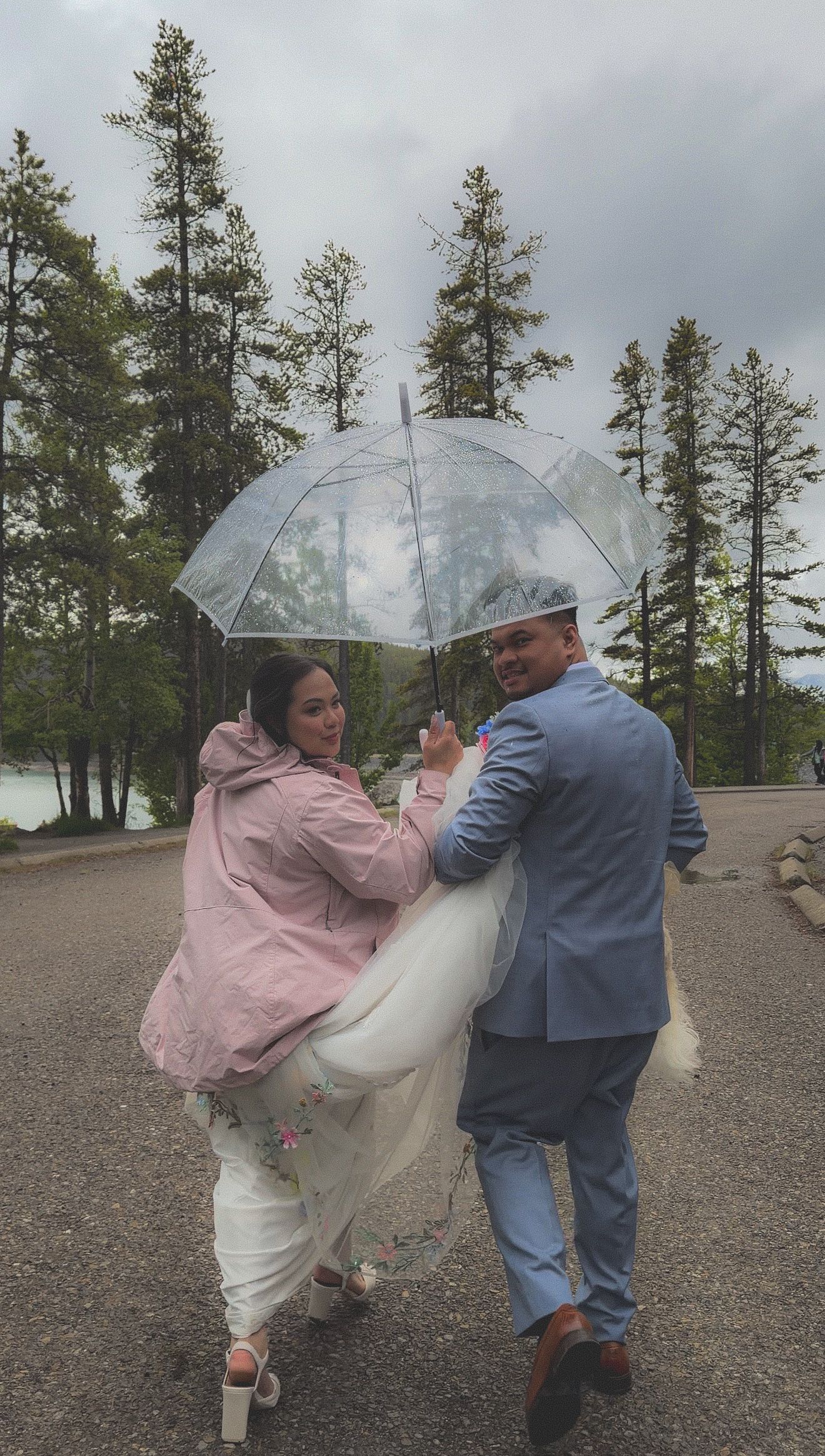 Newlywed couple sharing a joyful moment under a clear bubble umbrella in the rain near a forest.