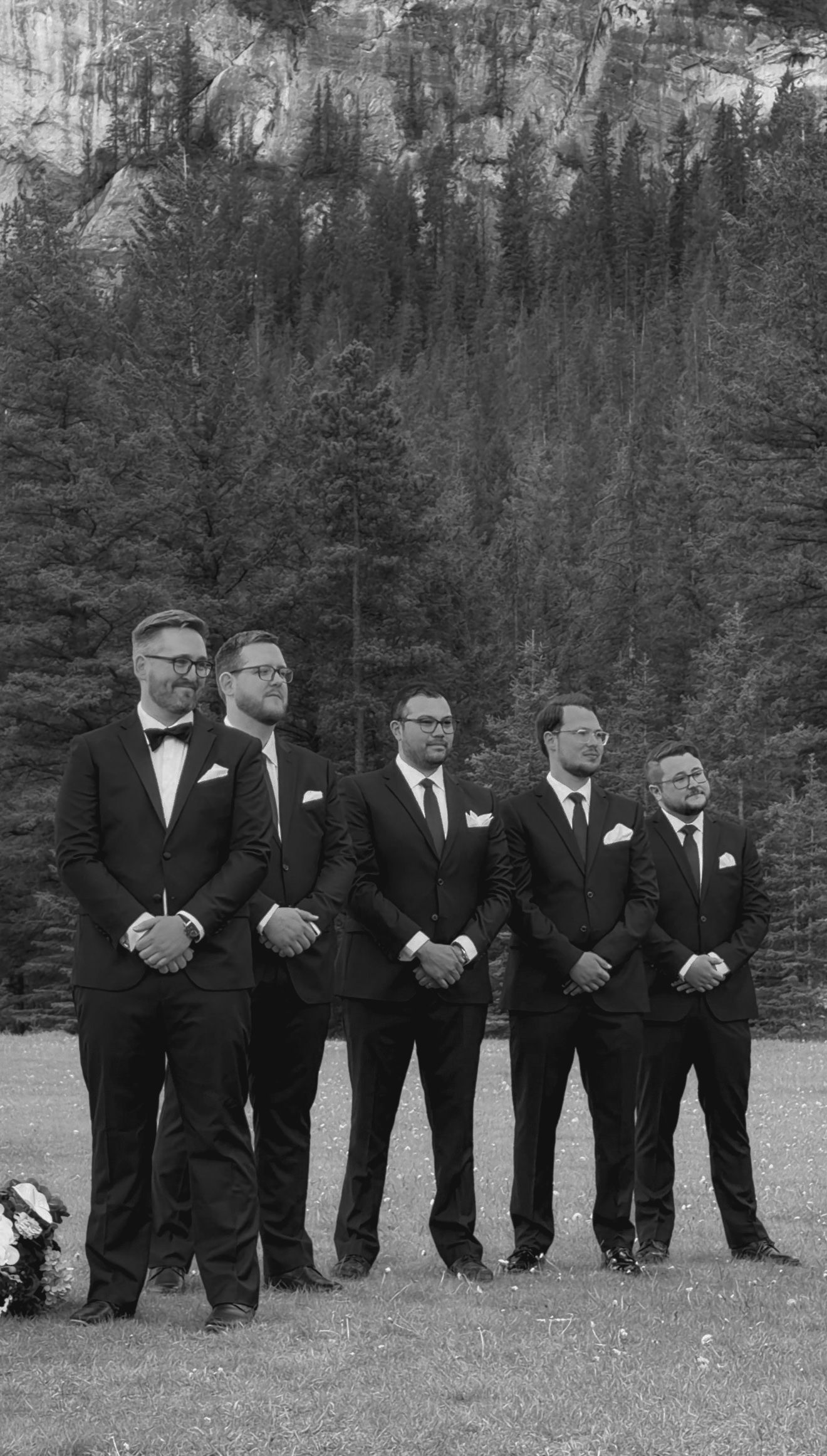 Five groomsmen in black tuxedos stand outdoors at a wedding ceremony with pine trees and rocky cliffs behind them.