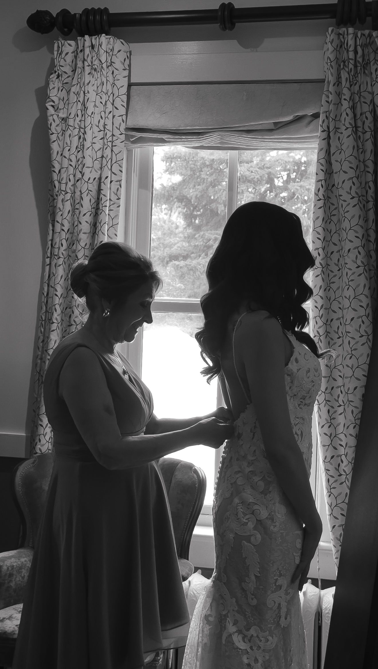 Black and white photo of mother helping bride button wedding dress by window.