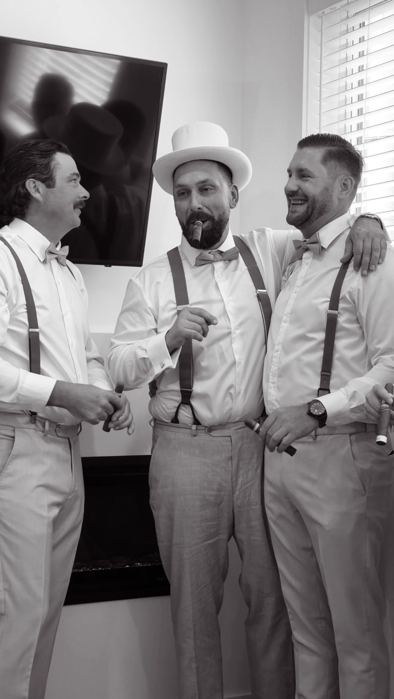 Three groomsmen in suspenders and bow ties laughing while smoking cigars before wedding ceremony.