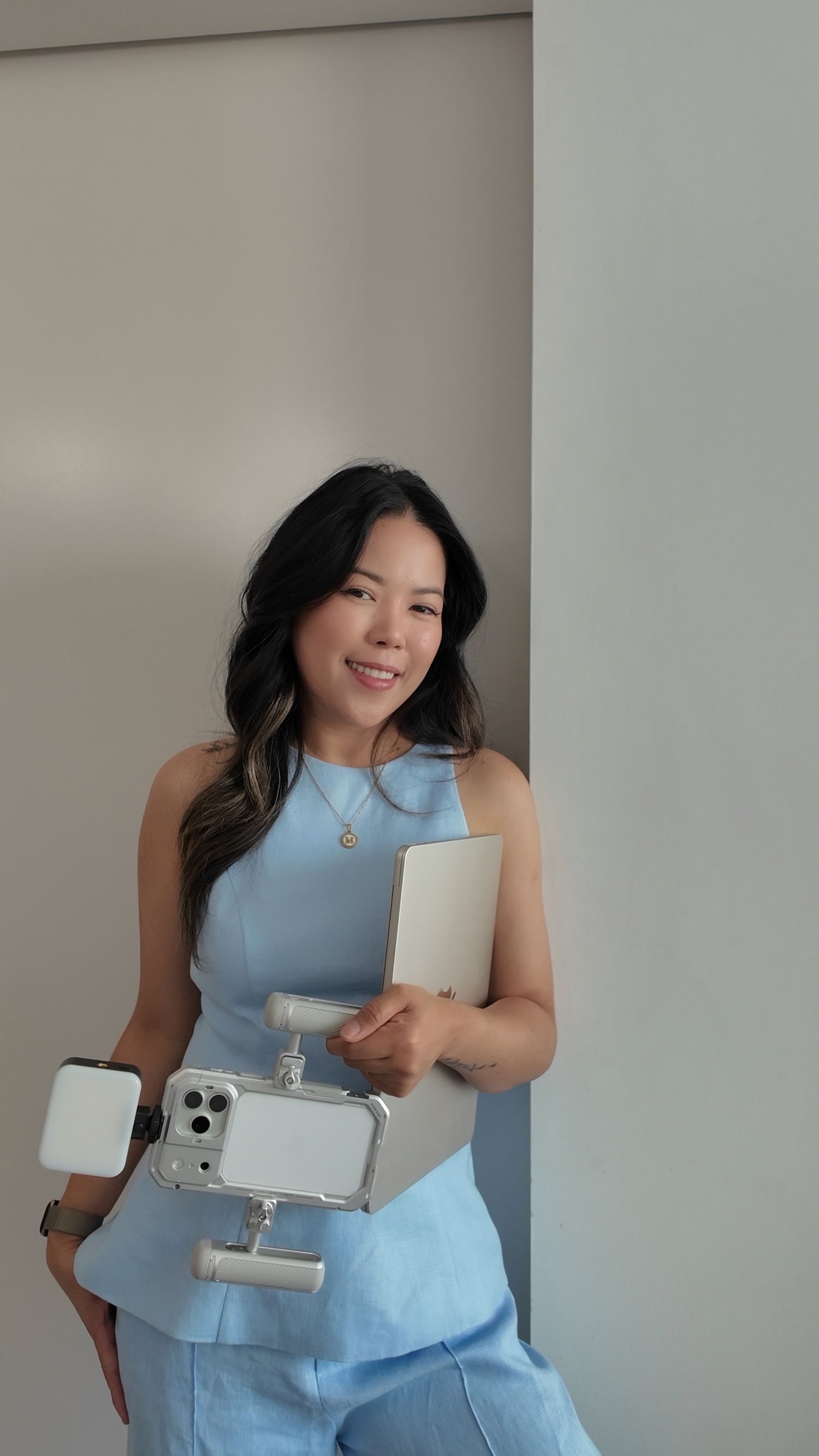 Woman in blue outfit holding laptop and phone gimbal stabilizer, smiling confidently against neutral background.