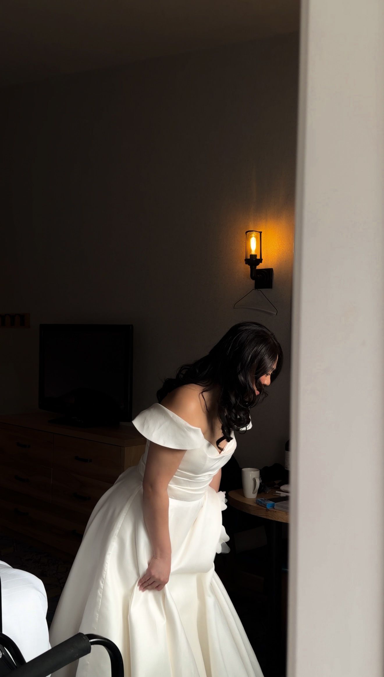 Bride in off-shoulder white wedding gown adjusting her dress in a warmly lit hotel room before ceremony.