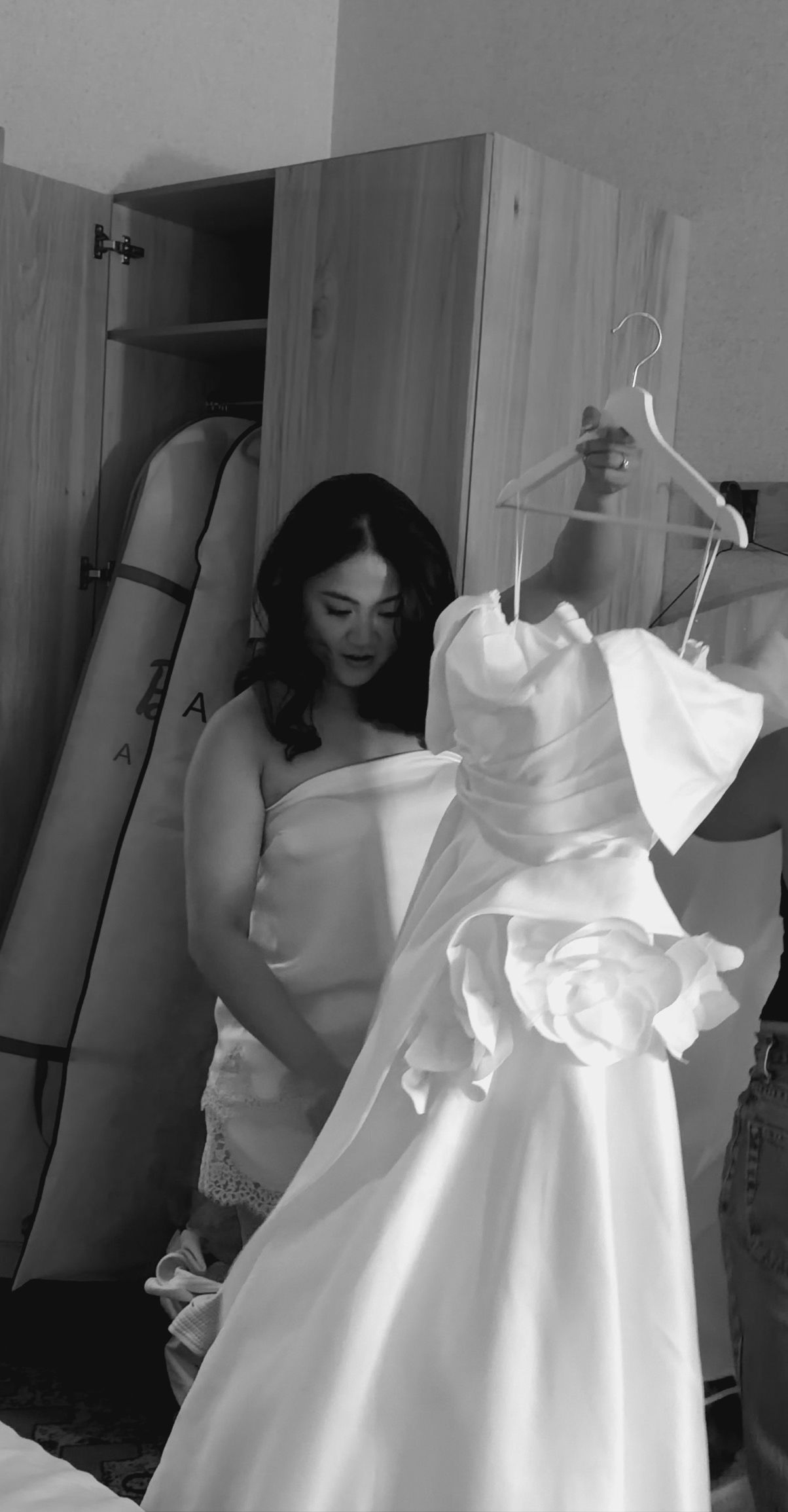 Black and white photo of bride admiring her strapless ruffled white wedding gown on a hanger indoors.