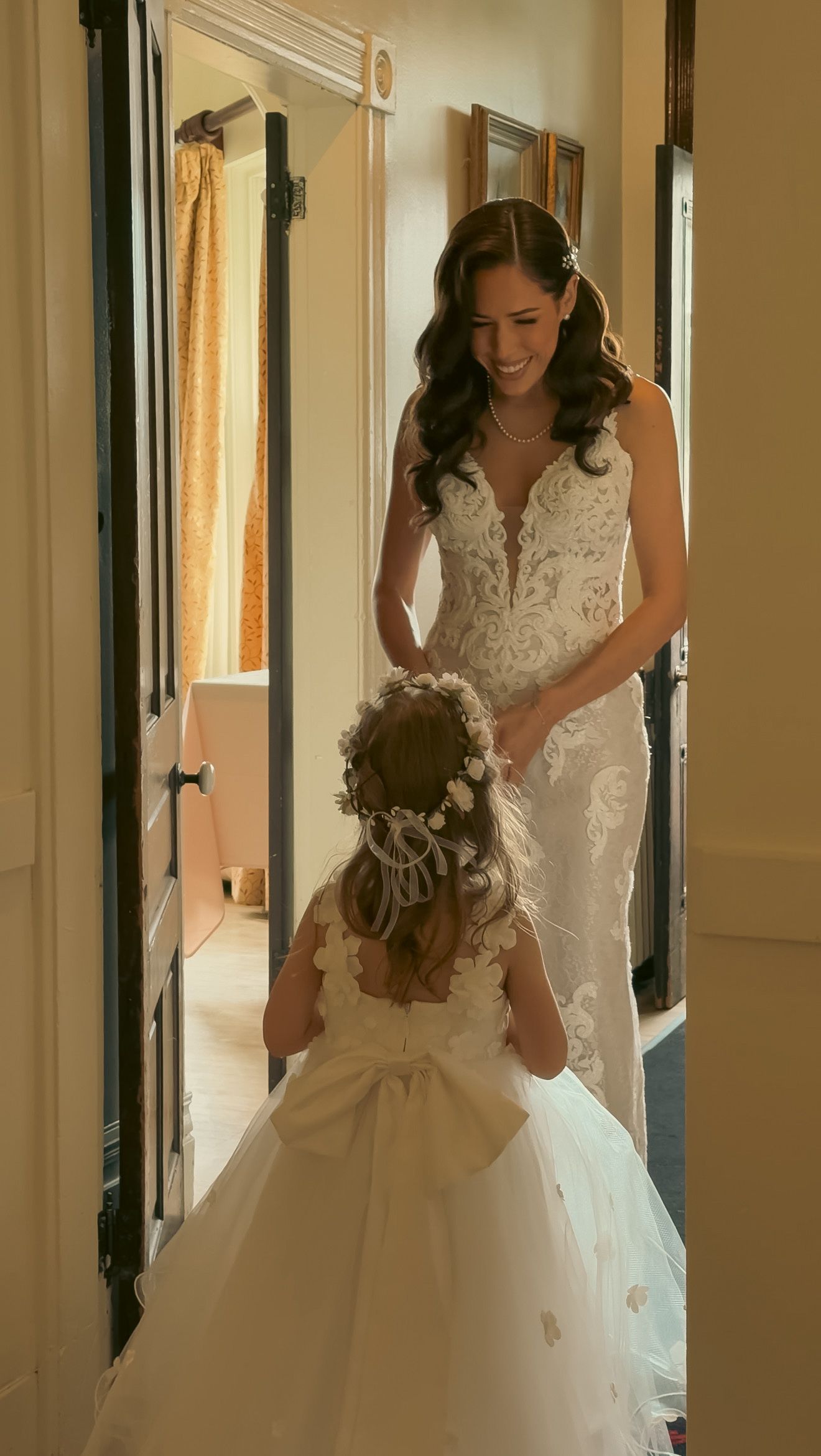 Radiant bride in lace wedding gown smiles warmly at flower girl in white dress.
