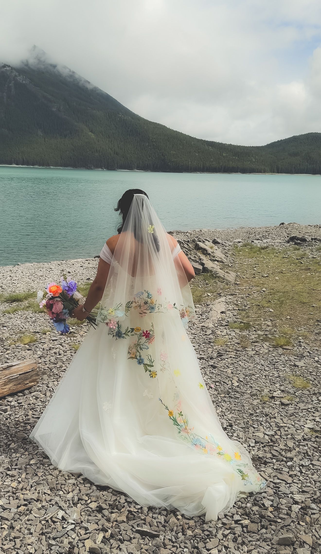 Bride in white embroidered gown with floral veil holding colorful bouquet by turquoise mountain lake.