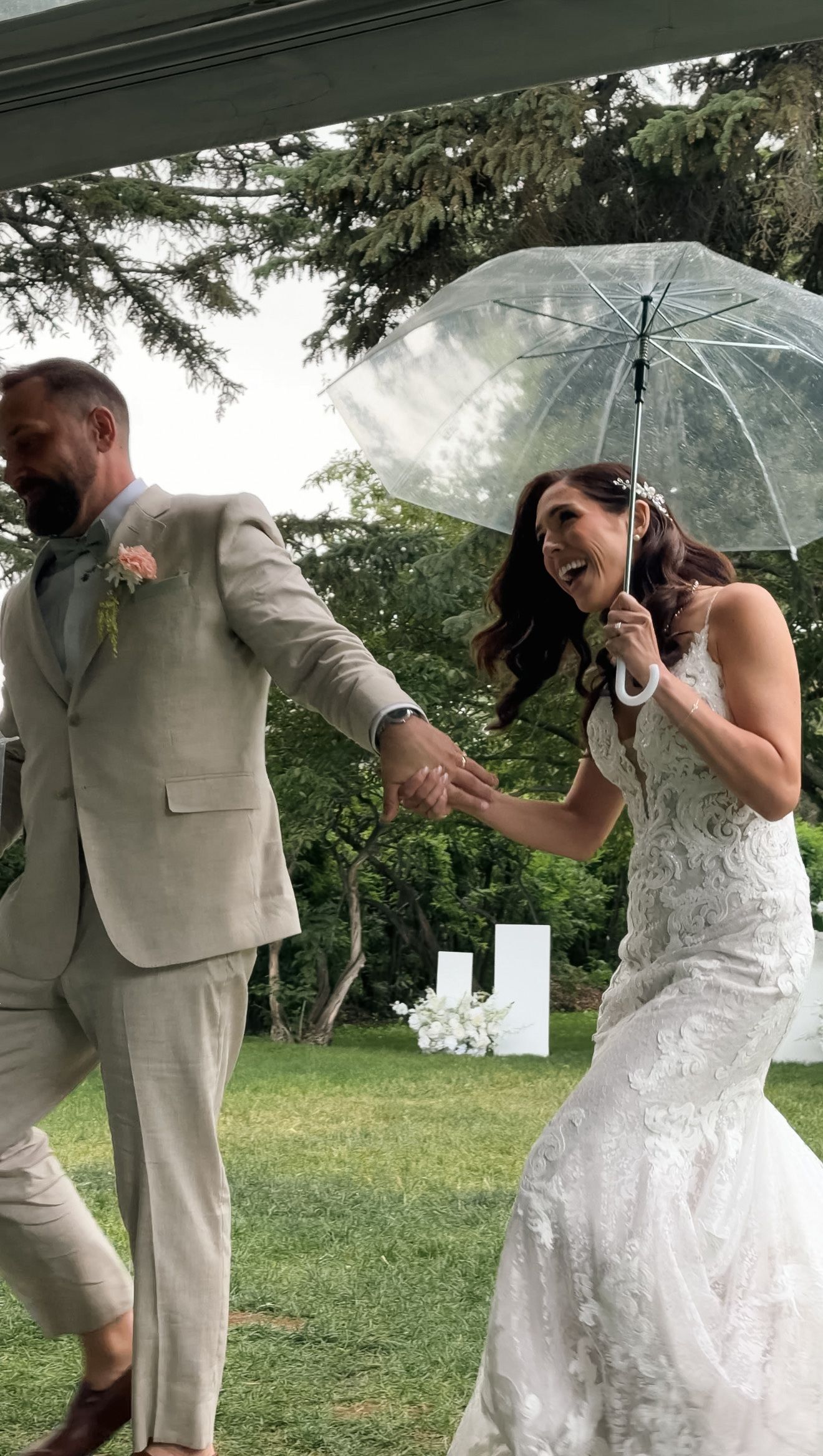 Laughing bride in lace gown holds clear umbrella while groom in tan suit leads her through rainy outdoor wedding.