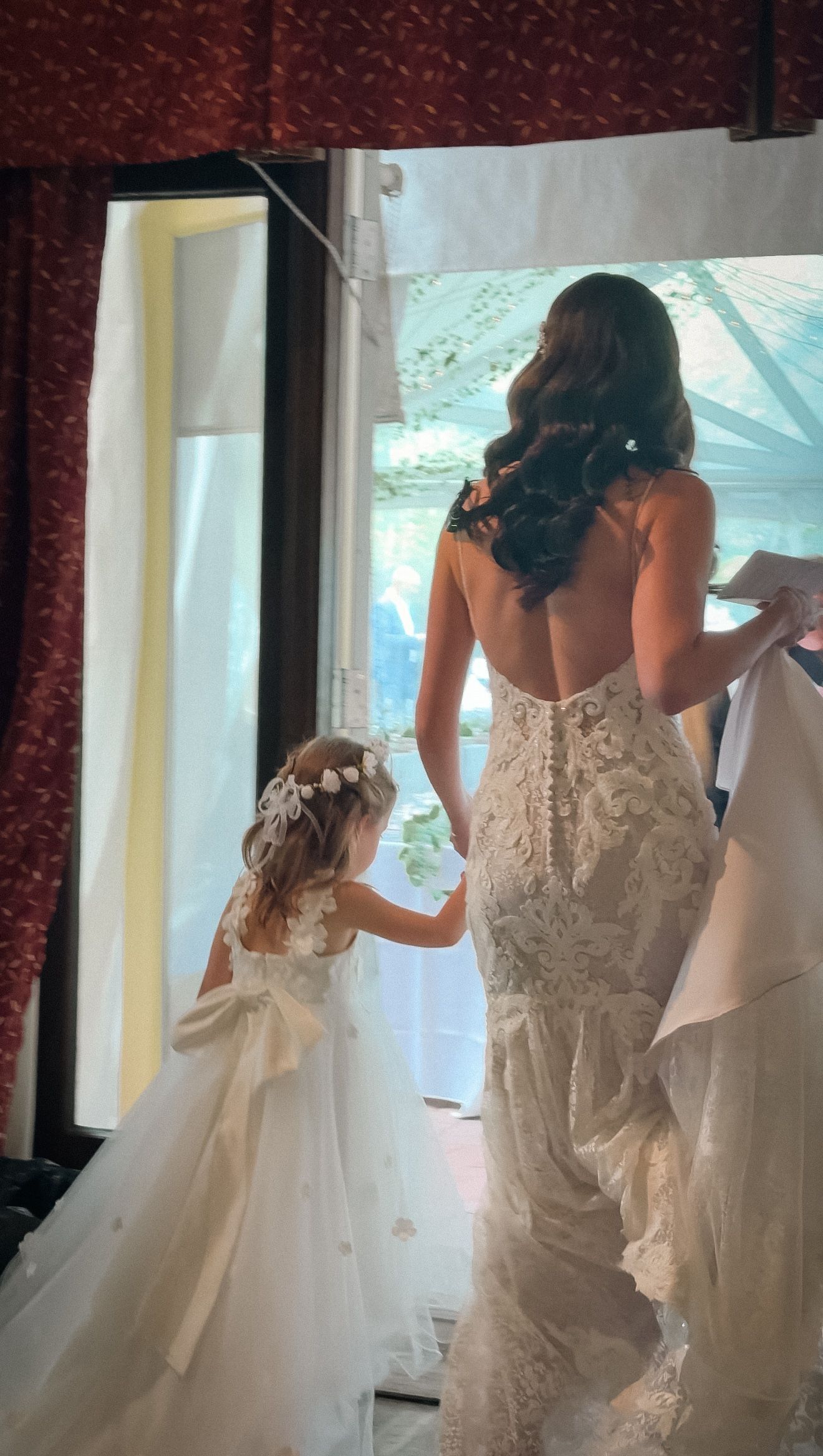 Bride in backless lace gown stands with adorable flower girl looking out window before ceremony.